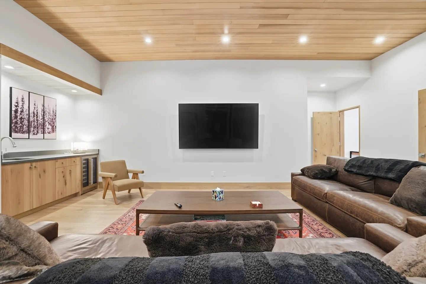 Living room with a wall-mounted TV, leather sofa, wood ceiling, wet bar, and large coffee table over an area rug.