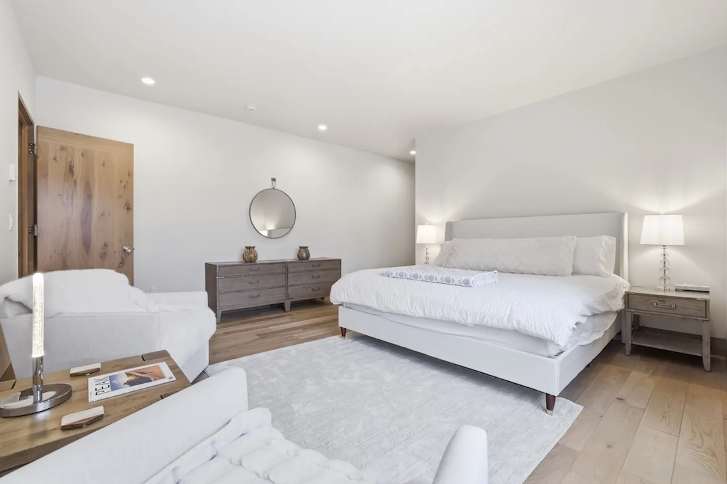 Bedroom with a white bed, light area rug, wood floor, rustic wooden dressers, and white accent armchairs.