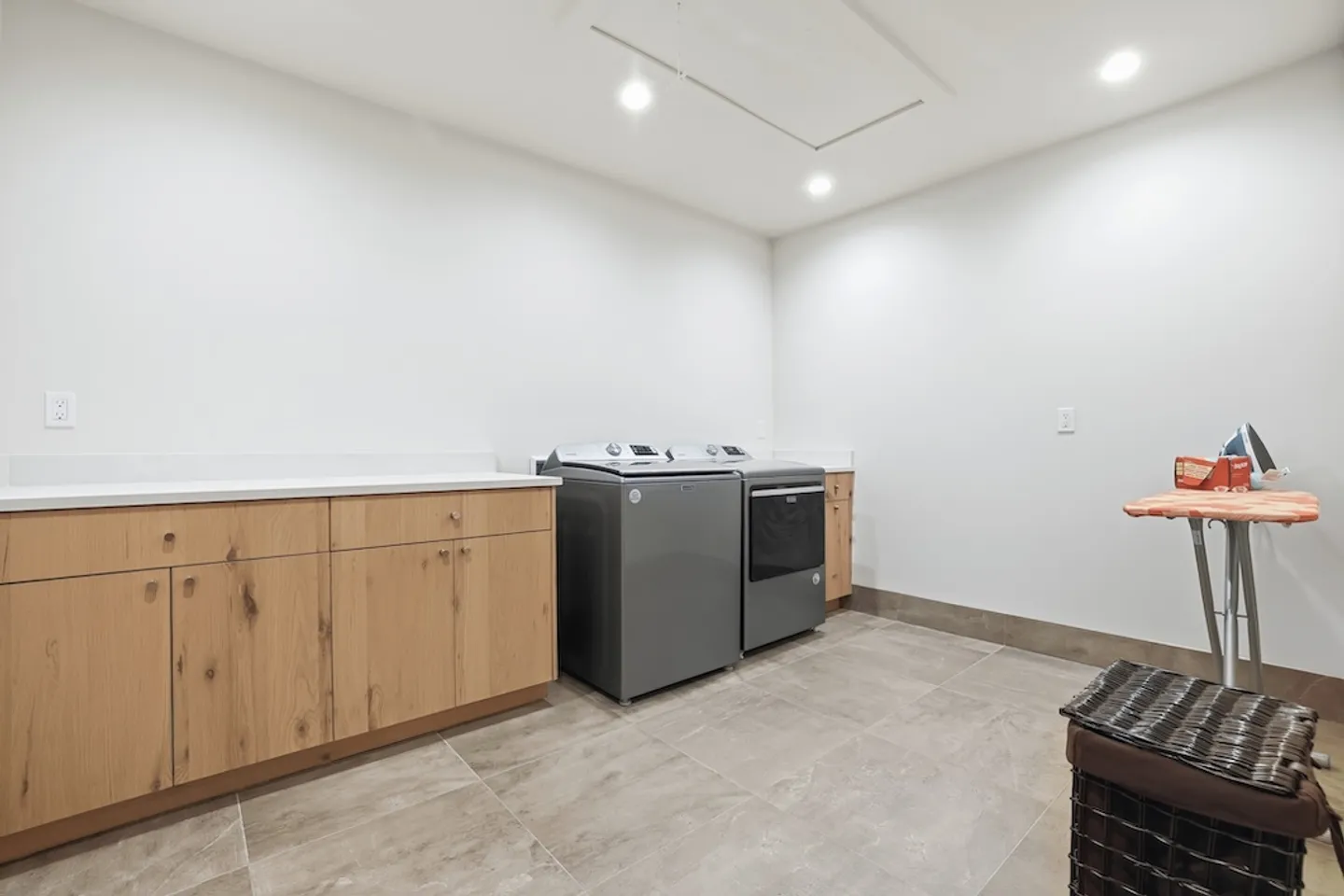 A brightly lit laundry room with a grey washer and dryer, wood-tone cabinets, and an ironing board.
