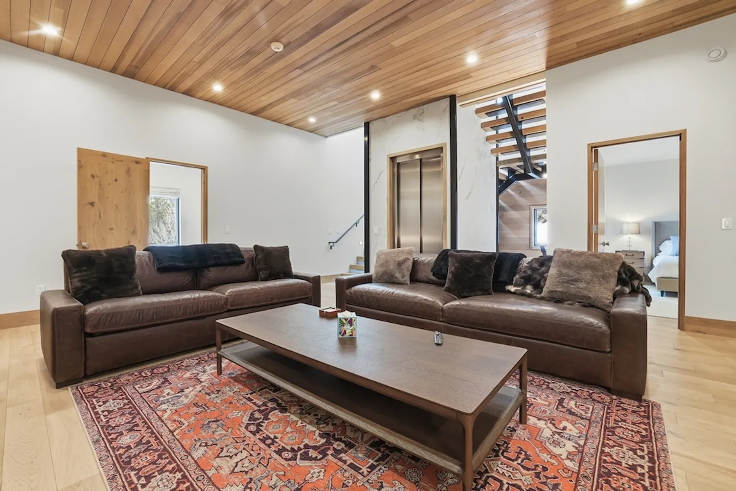 Living area featuring two brown leather sofas, a wood coffee table, an elevator, and a modern open staircase under a wood-plank ceiling.