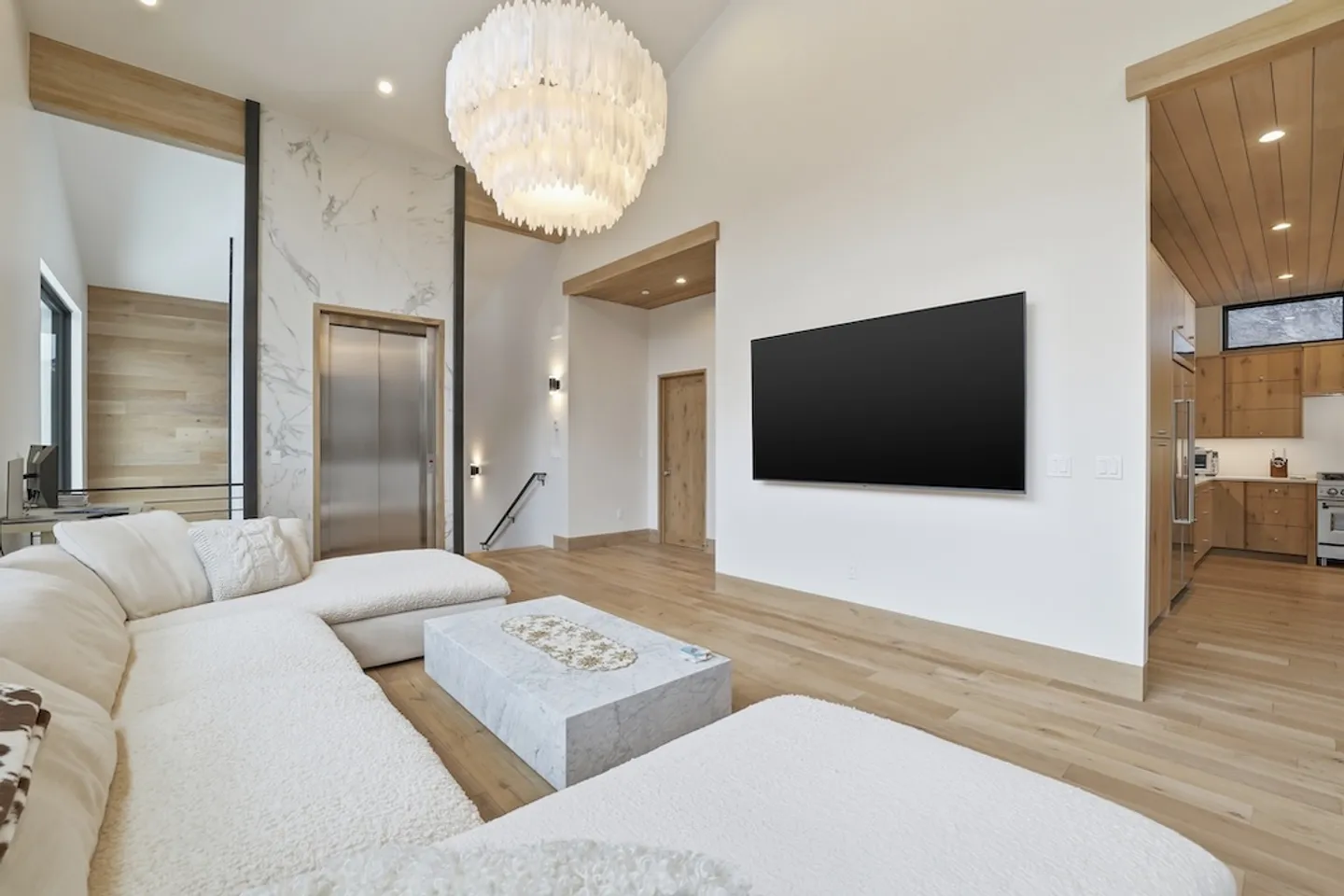 Open-concept living room featuring light wood floors, a sectional sofa, a marble coffee table, a wall-mounted TV, an elevator, and a modern chandelier.