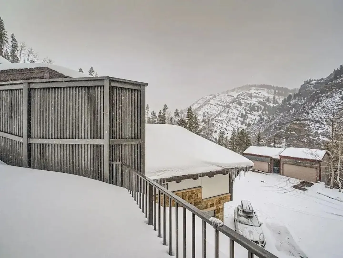 Winter mountain view from a snow-covered deck railing looking toward rooftops and a snowy hillside.