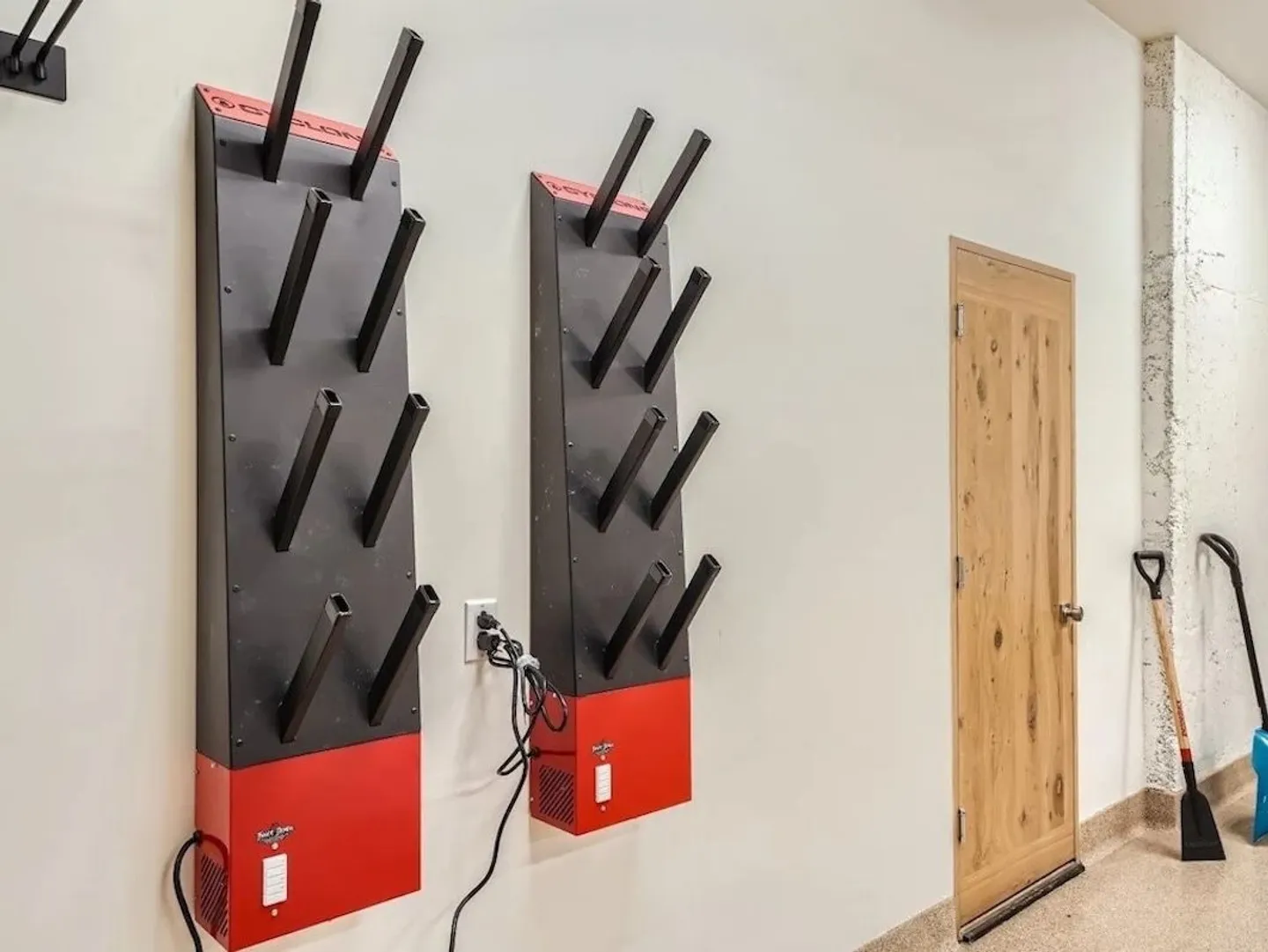 Two wall-mounted electric boot dryers with angled pegs plugged into an outlet next to a light wood door.