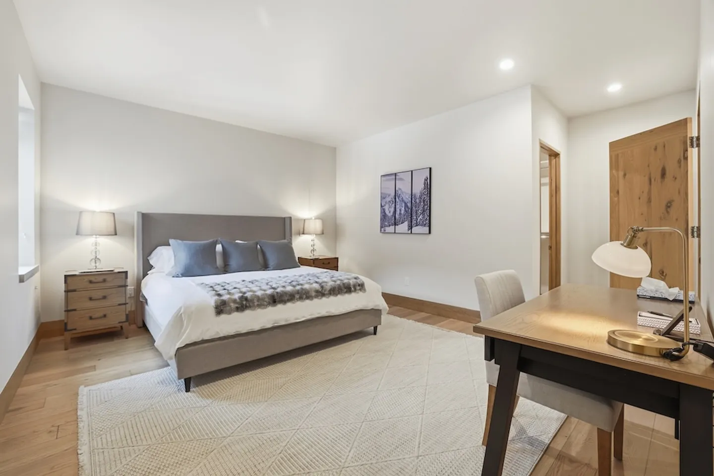 Bedroom with an upholstered bed, nightstands with lamps, a patterned area rug on wood flooring, and a desk with a lamp in the foreground.