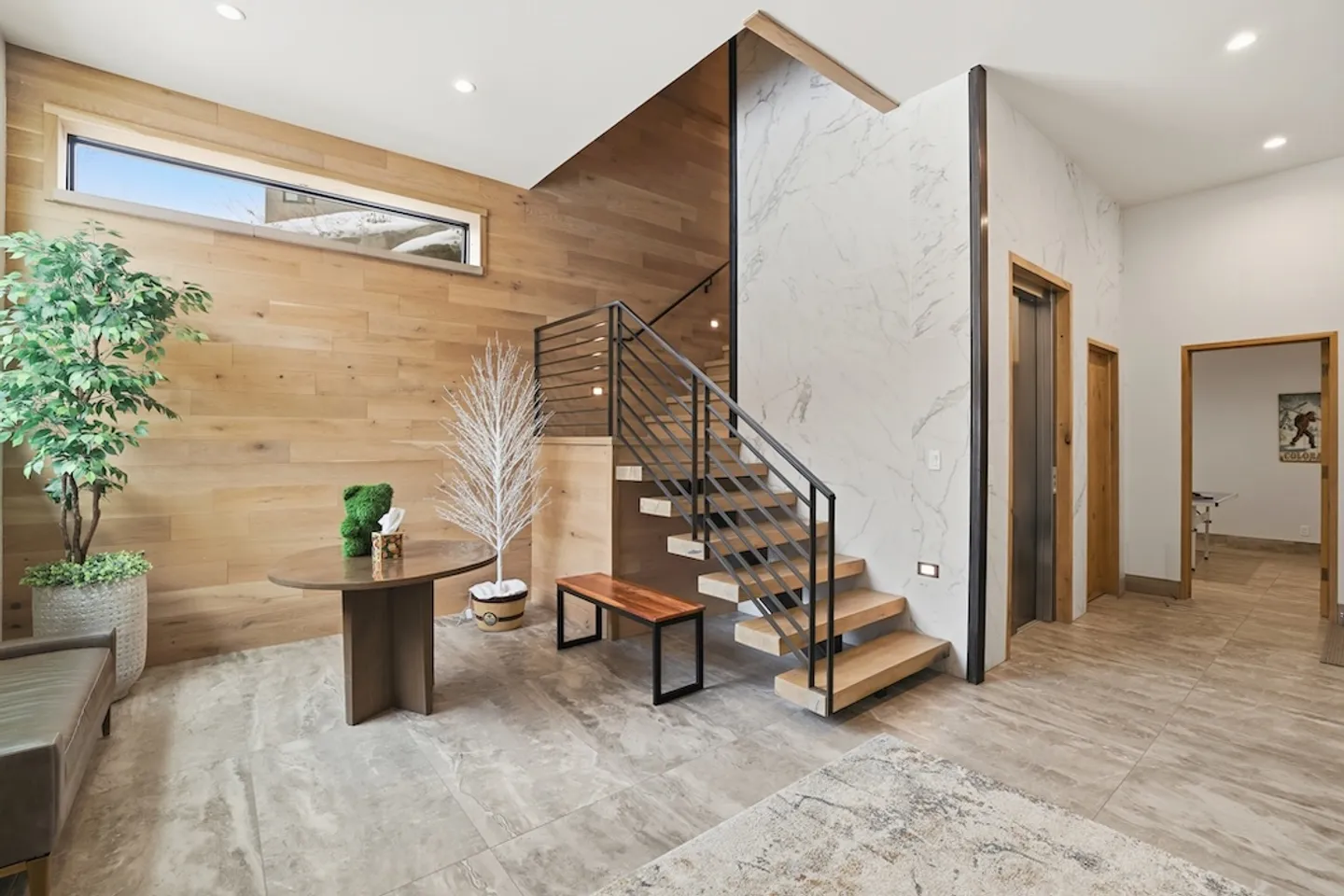 A modern entryway with a floating wood-tread staircase, wood-paneled accent wall, marble-look accent wall, and an elevator door.