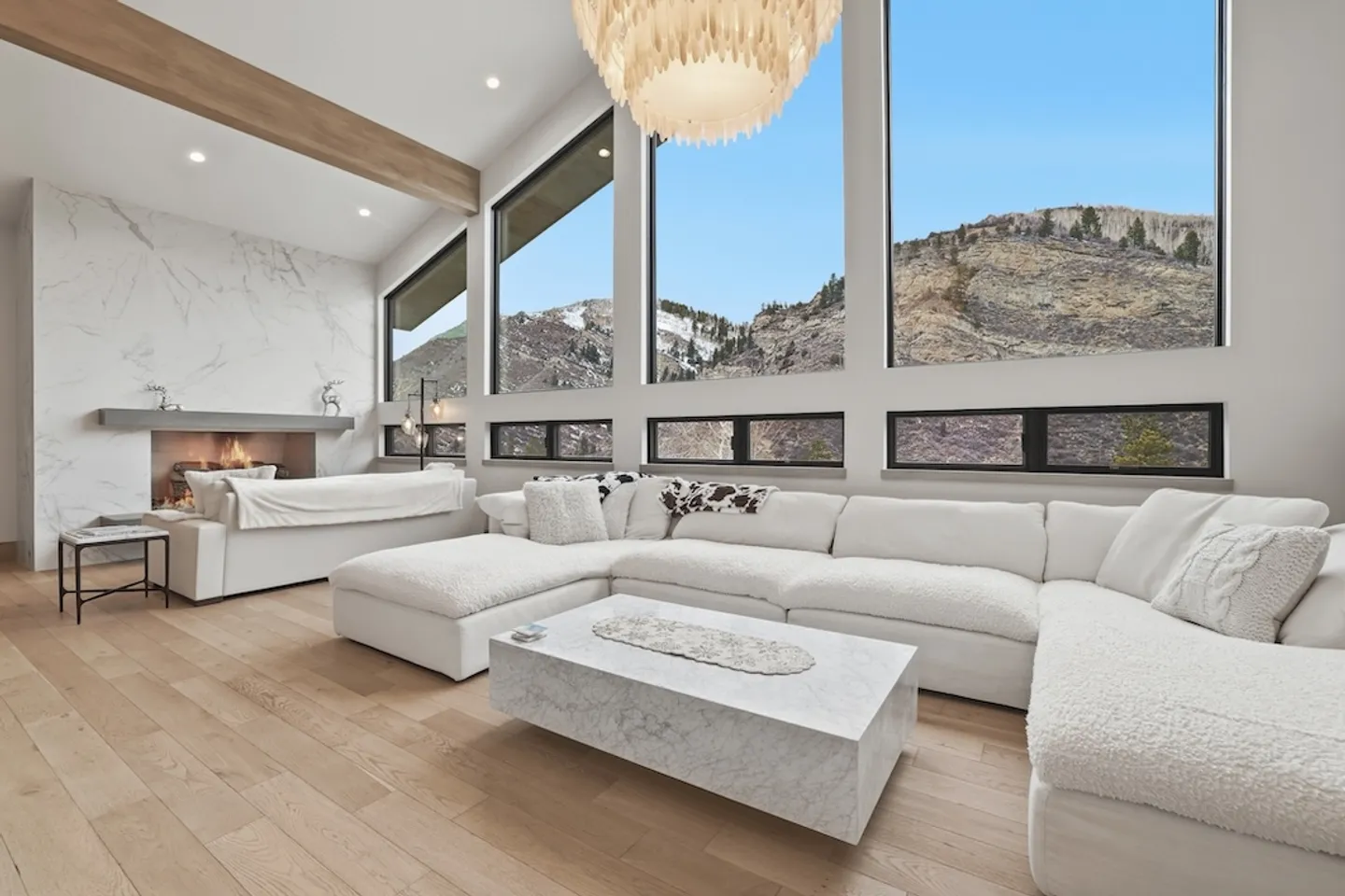 Bright modern living room with large windows showing mountain views, a white sectional sofa, marble coffee table, and a fireplace.