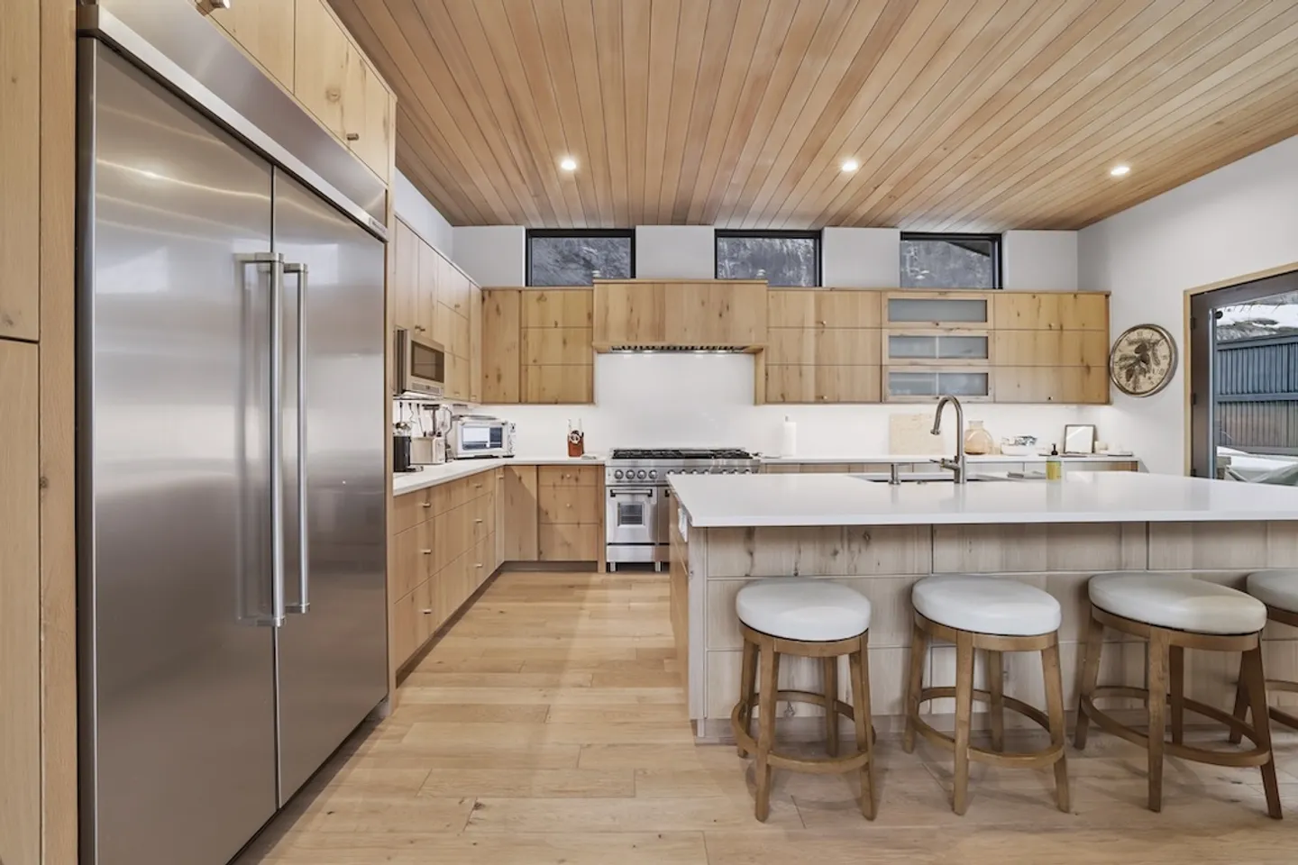A modern kitchen with light wood cabinets, wood-plank ceiling, kitchen island with barstools, and stainless steel appliances.