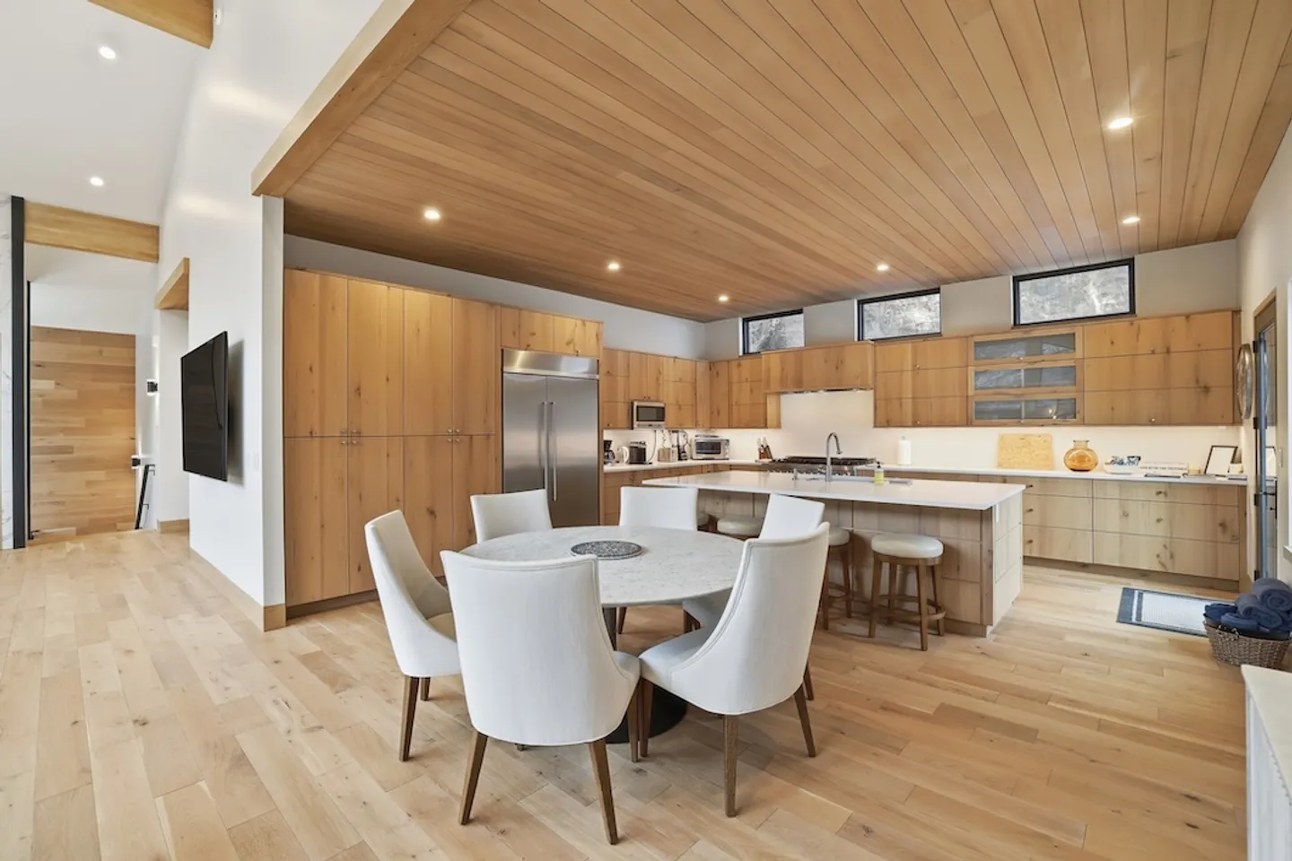 Open-concept kitchen and dining area featuring light wood cabinetry, wood-paneled ceiling, a round dining table with white chairs, and a central kitchen island.