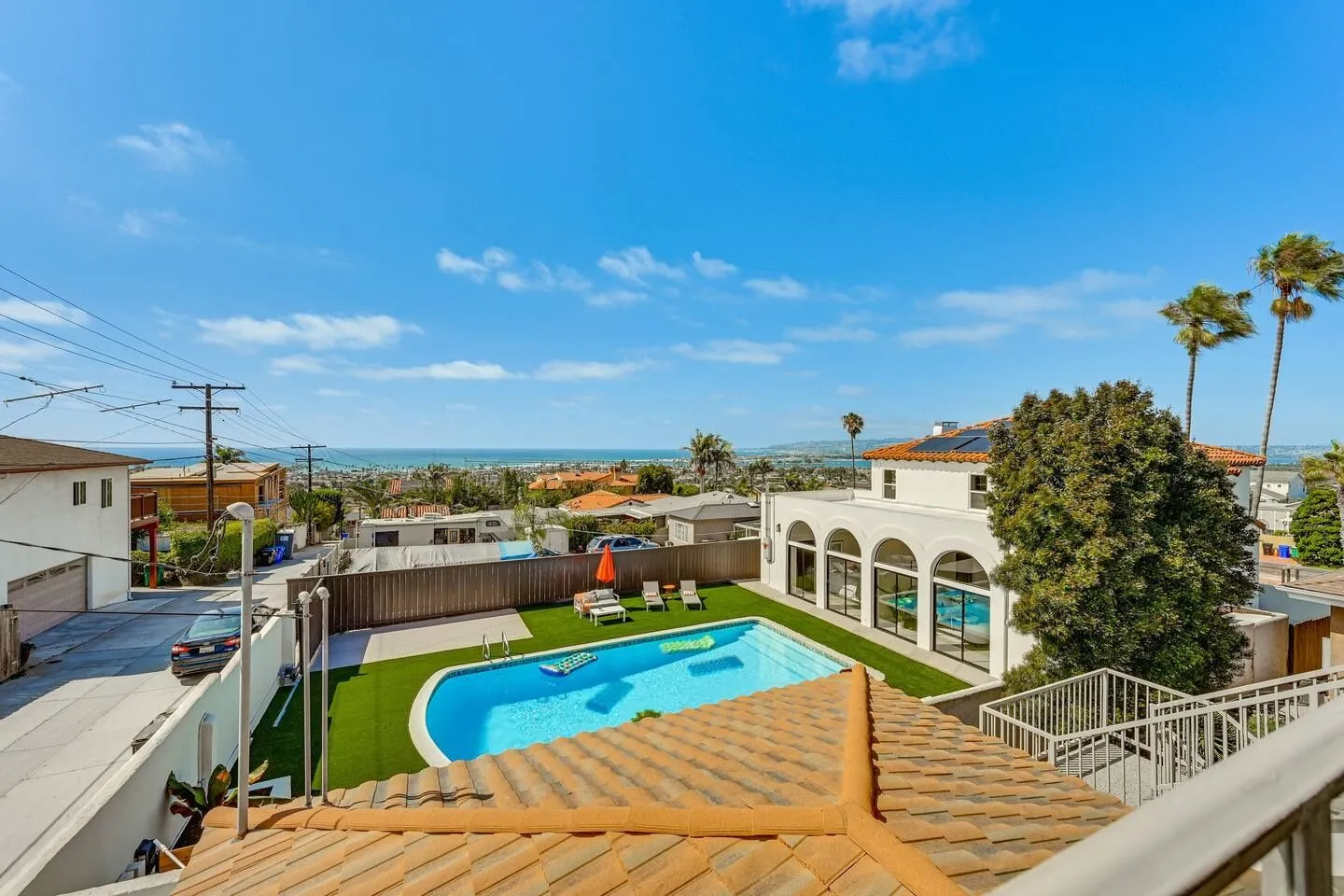 High-angle view of a backyard with a swimming pool, lounge furniture, and views of the ocean in the distance under a blue sky.