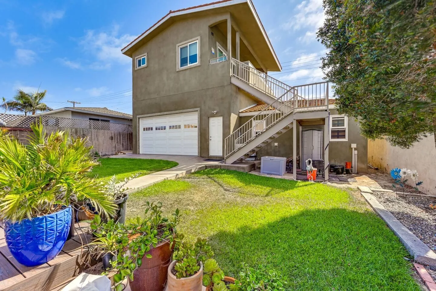 Backyard lawn and patio leading to a two-story building with a garage, exterior staircase, and upper-level balcony under a blue sky.