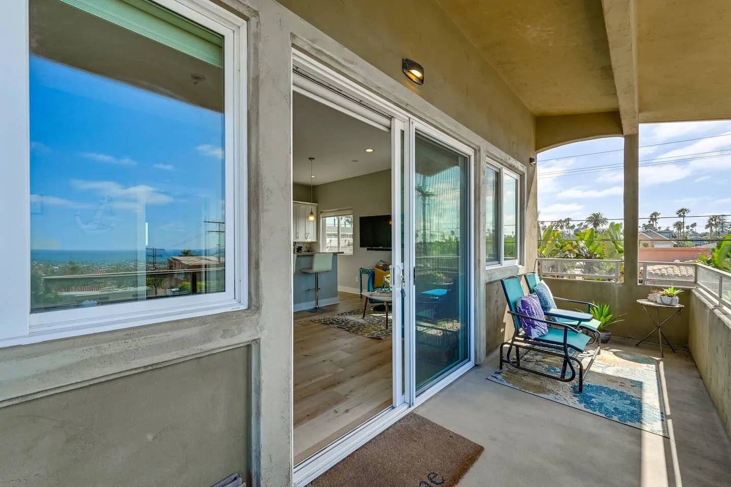 A covered balcony with outdoor chairs looking out toward palm trees and the ocean, connected by sliding glass doors to the living area.
