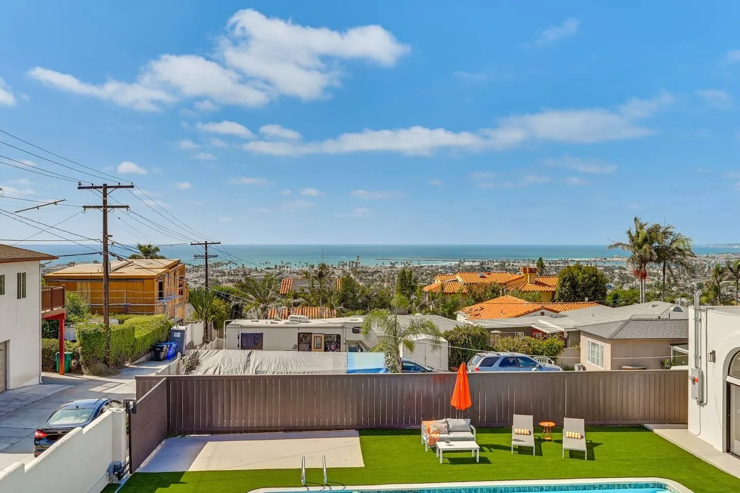 High-angle view of a backyard with artificial turf, outdoor seating, and a pool edge, overlooking a neighborhood and the ocean in the distance.