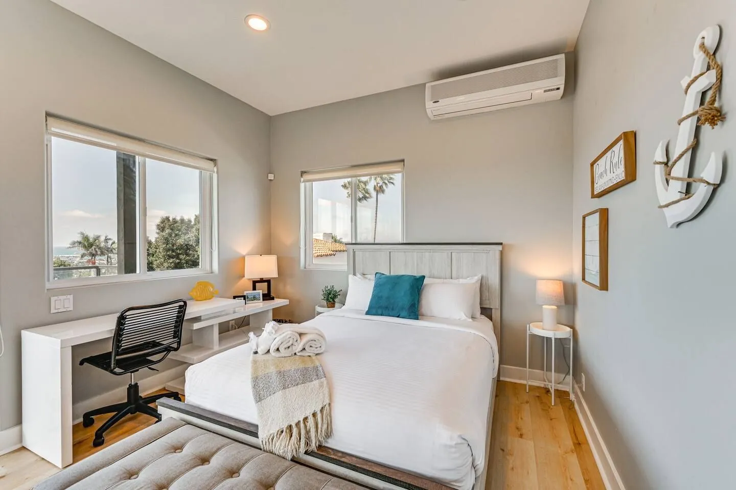 Bedroom with a made bed, wall-mounted air conditioner, light wood floors, a built-in desk, and windows showing trees and sky.