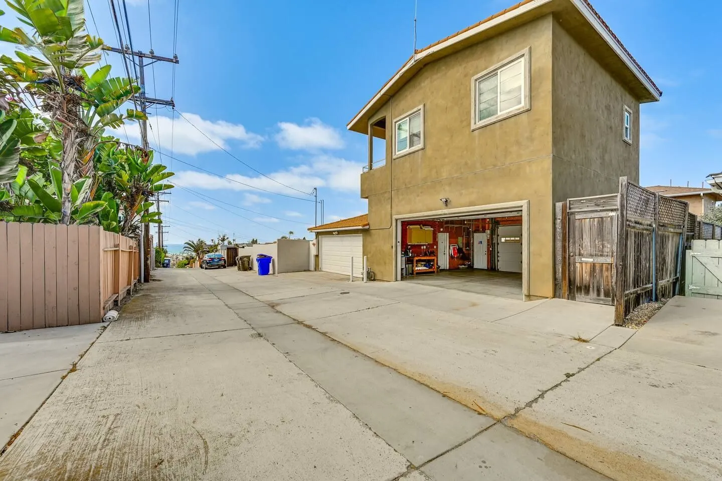 Exterior view of an alleyway leading down towards the ocean with a two-story home, an open garage, and fenced properties.