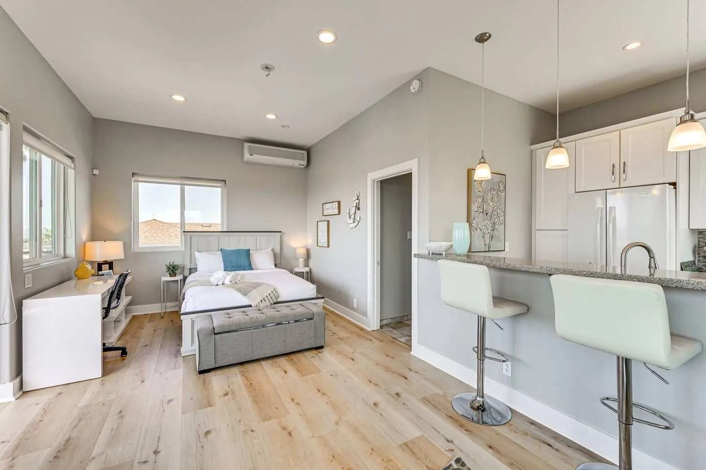 Open-concept studio apartment featuring a bed, workspace desk, breakfast bar with barstools, and kitchen area with light wood flooring.