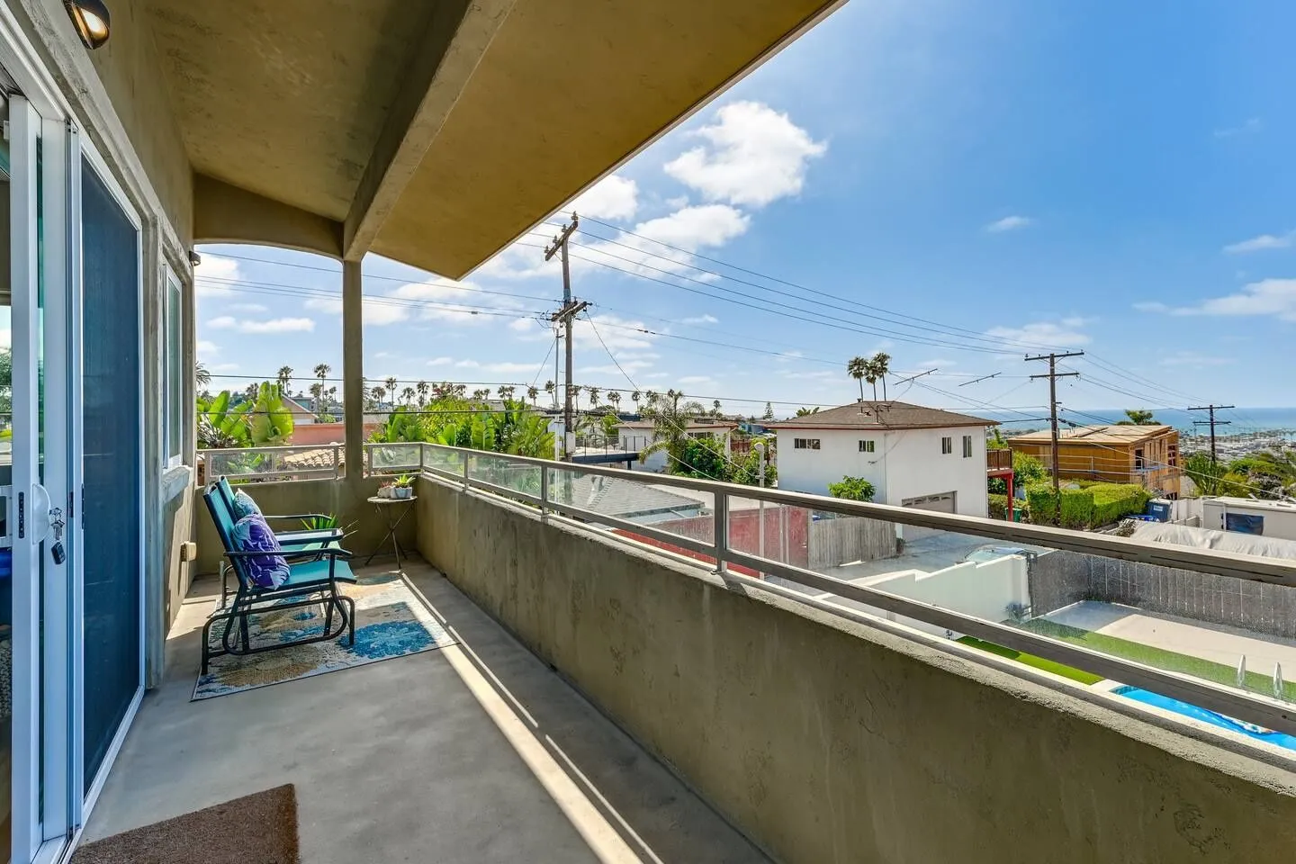 Covered outdoor balcony with seating area and glass railing overlooking a neighborhood toward the ocean.