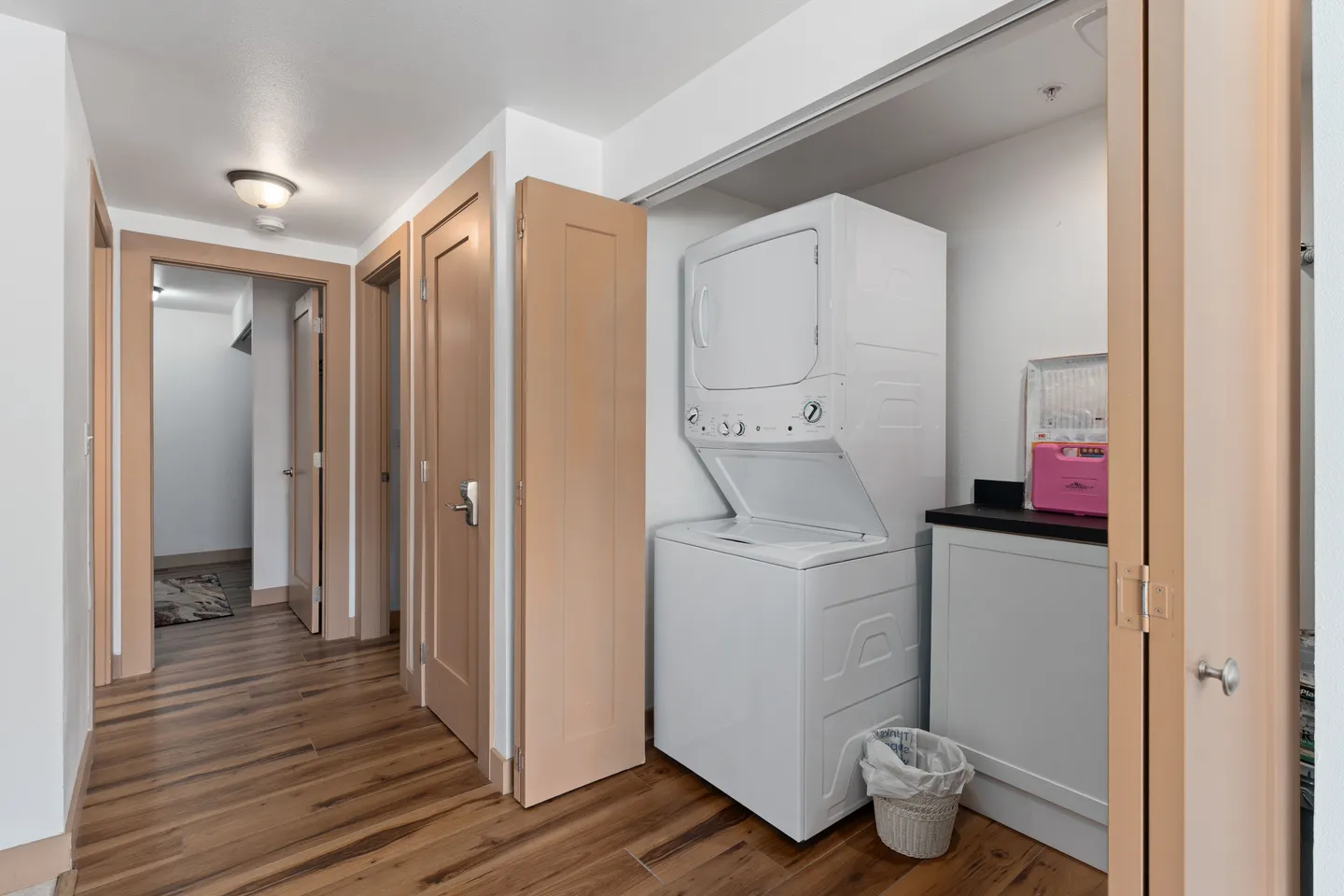 A hallway closet with bi-fold doors open to reveal a stacked washer and dryer unit, a small counter cabinet, and a wastebasket on wood-look flooring.