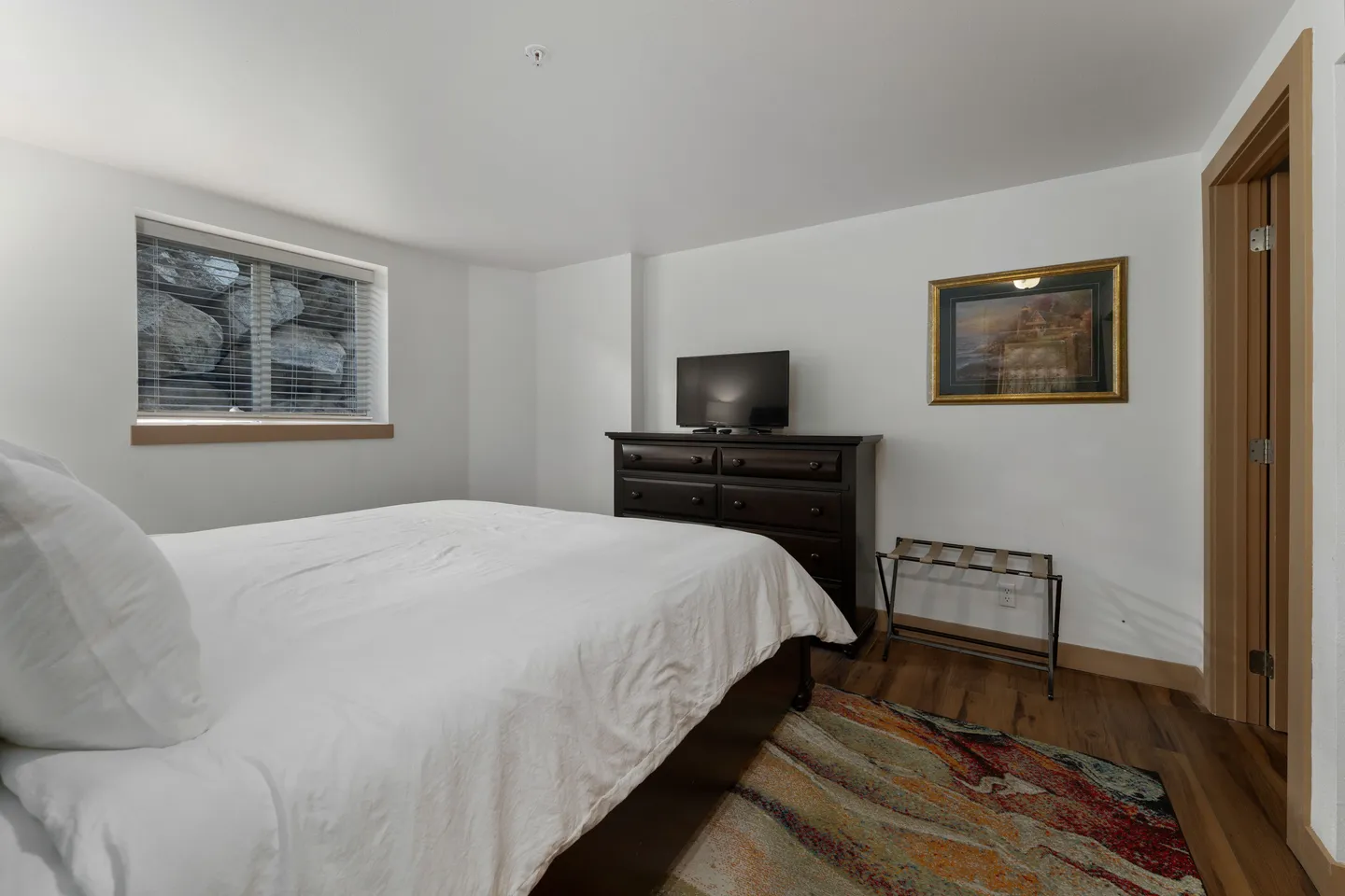 Bedroom with a bed made with white linens, a dark wood dresser with a TV, and a window with blinds overlooking a rock wall.
