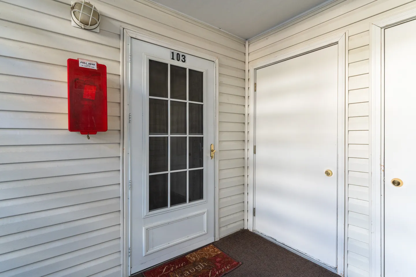 Exterior doorway to unit 103 with a paneled screen door and a red fire alarm pull station on siding.