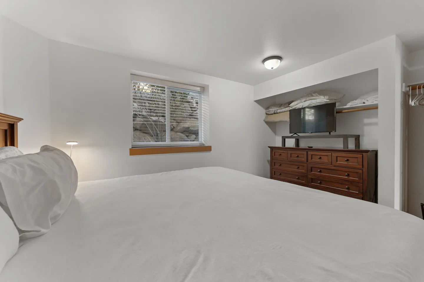 Bedroom with a bed dressed in white linens, a window with horizontal blinds, and a television on a wooden dresser.