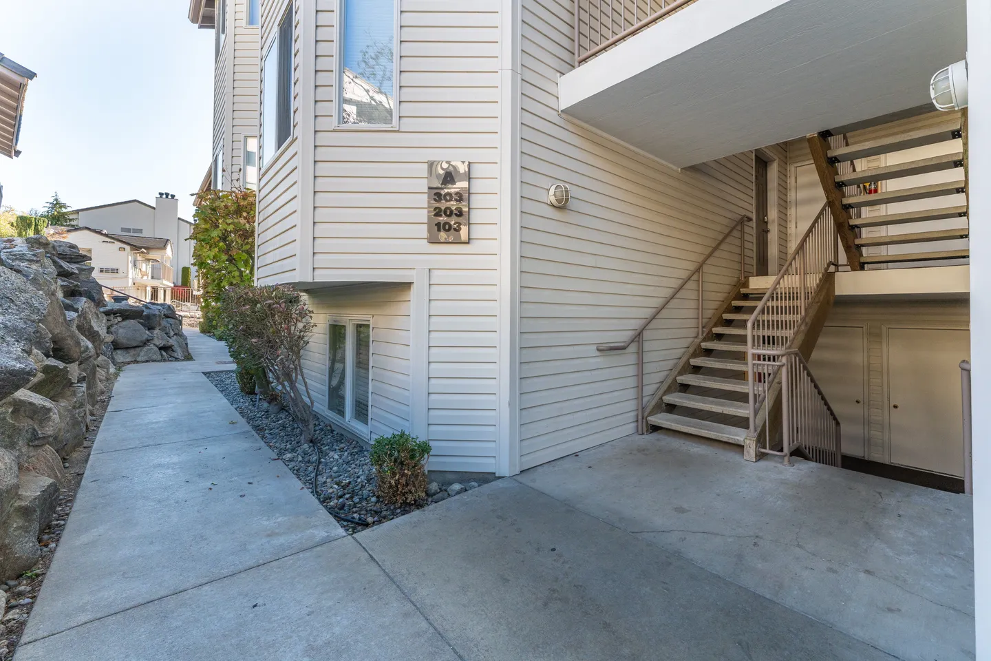 Exterior entryway of a multi-unit residential building with a concrete walkway, rock retaining wall, and stairs leading to upper levels.