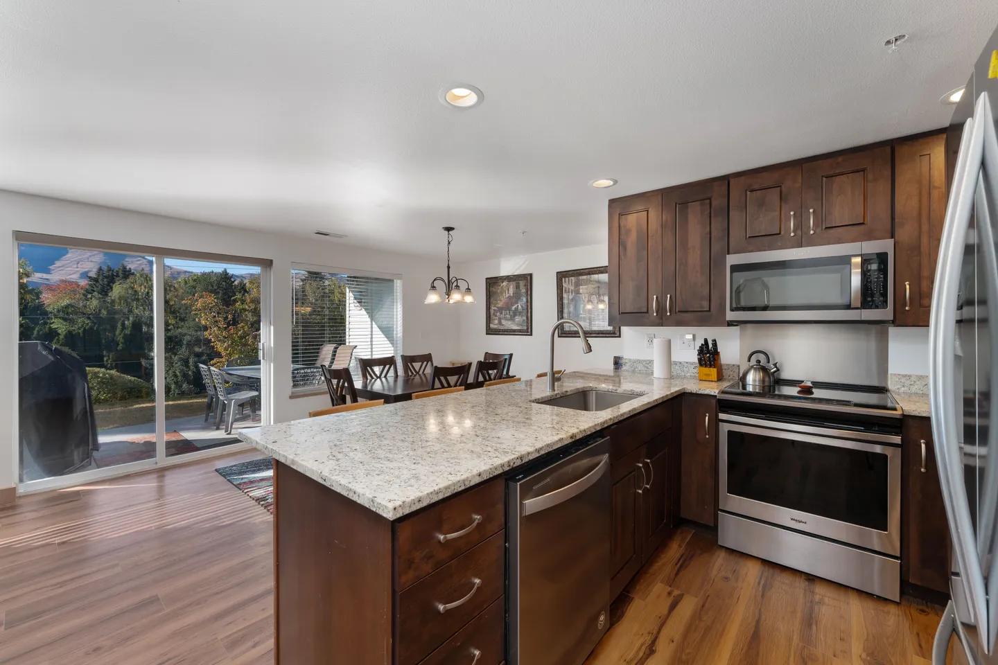 Open-concept kitchen with dark wood cabinets, speckled stone countertops, stainless steel appliances, and a dining area beside sliding glass patio doors.