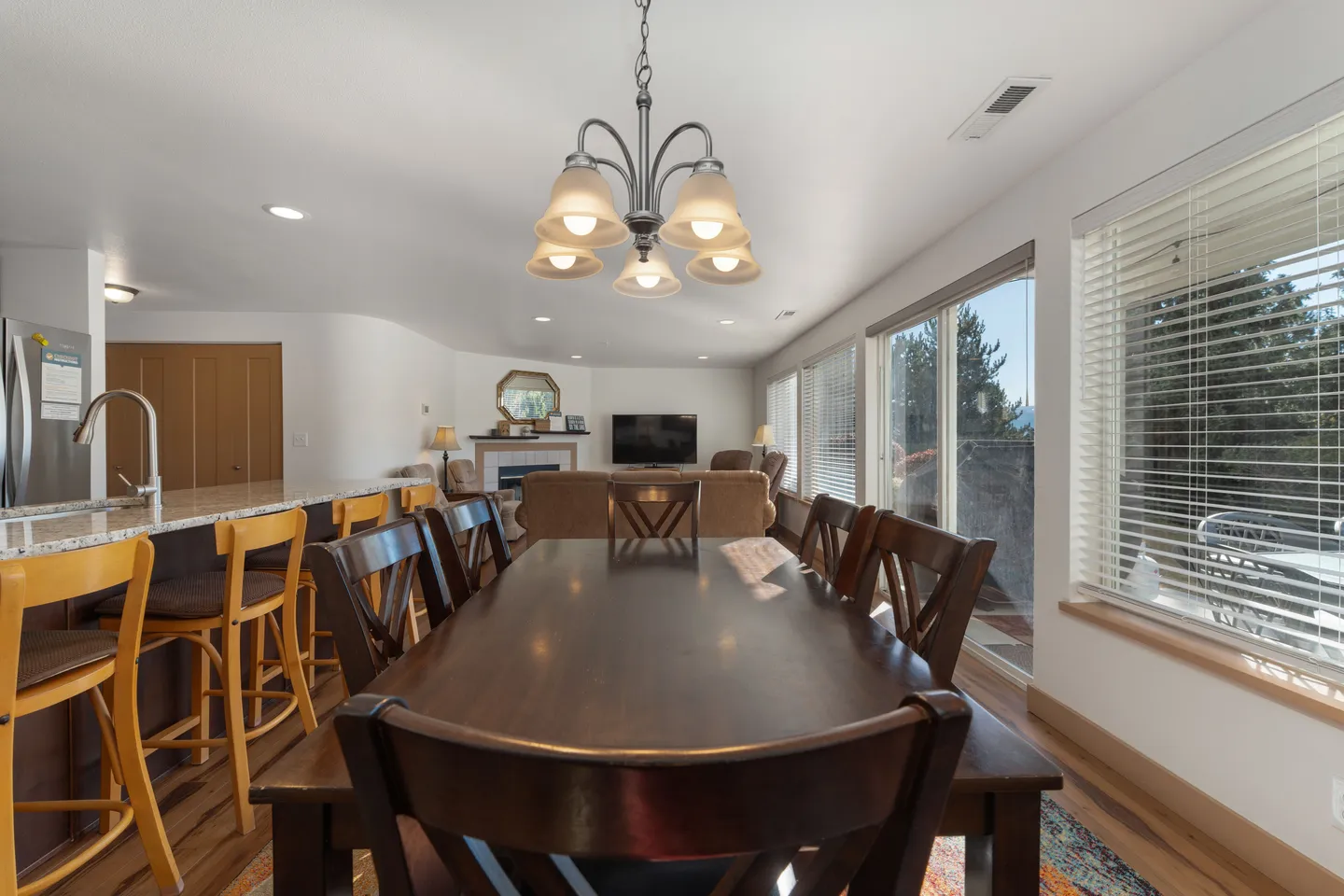 Dining table and chairs under a chandelier adjacent to a kitchen counter and living room area.