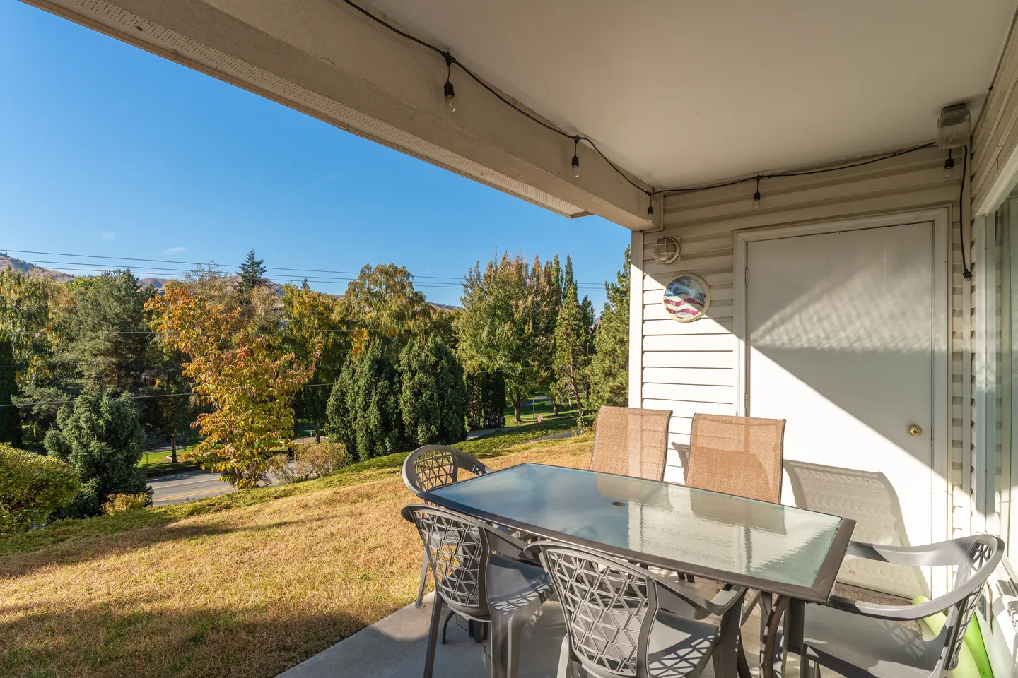Covered patio with an outdoor dining table and chairs overlooking a lawn, trees, and distant hills under a clear blue sky.