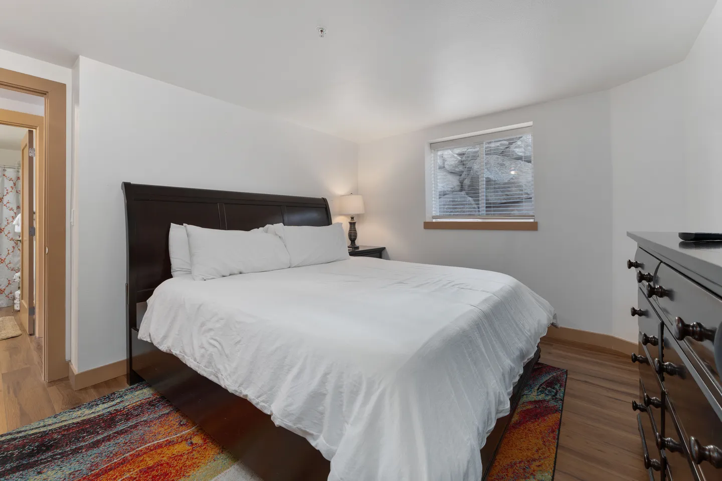 Bedroom with a dark wood sleigh bed, white bedding, wood-look flooring, a dresser, and a window with blinds showing a rocky retaining wall.