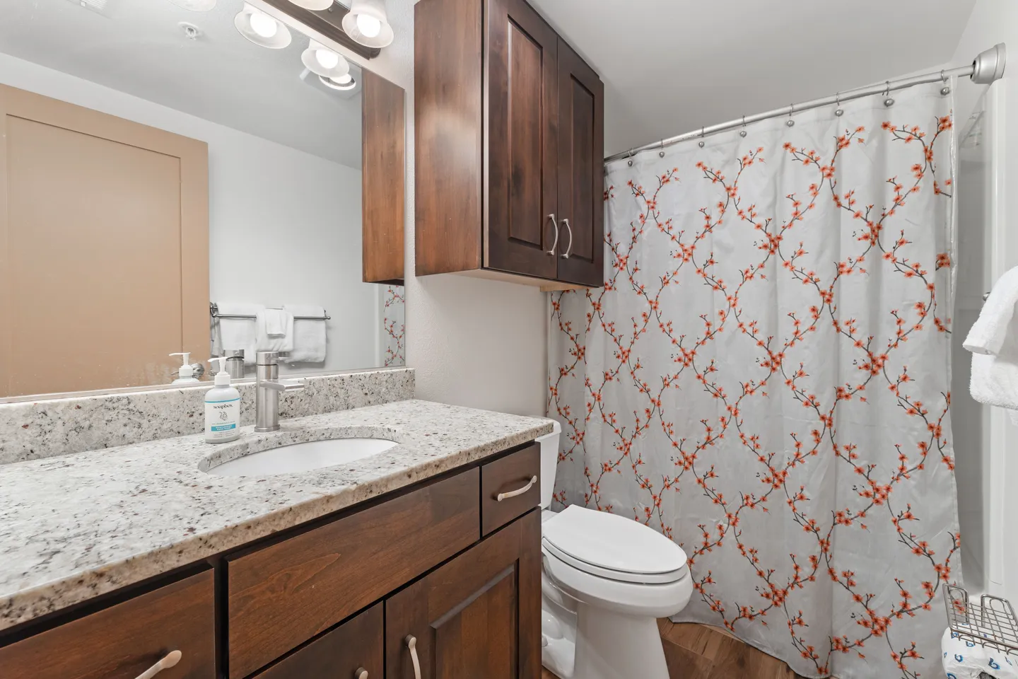 Bathroom featuring a dark wood vanity with speckled countertop, a toilet, an overhead cabinet, and a shower with a patterned curtain.