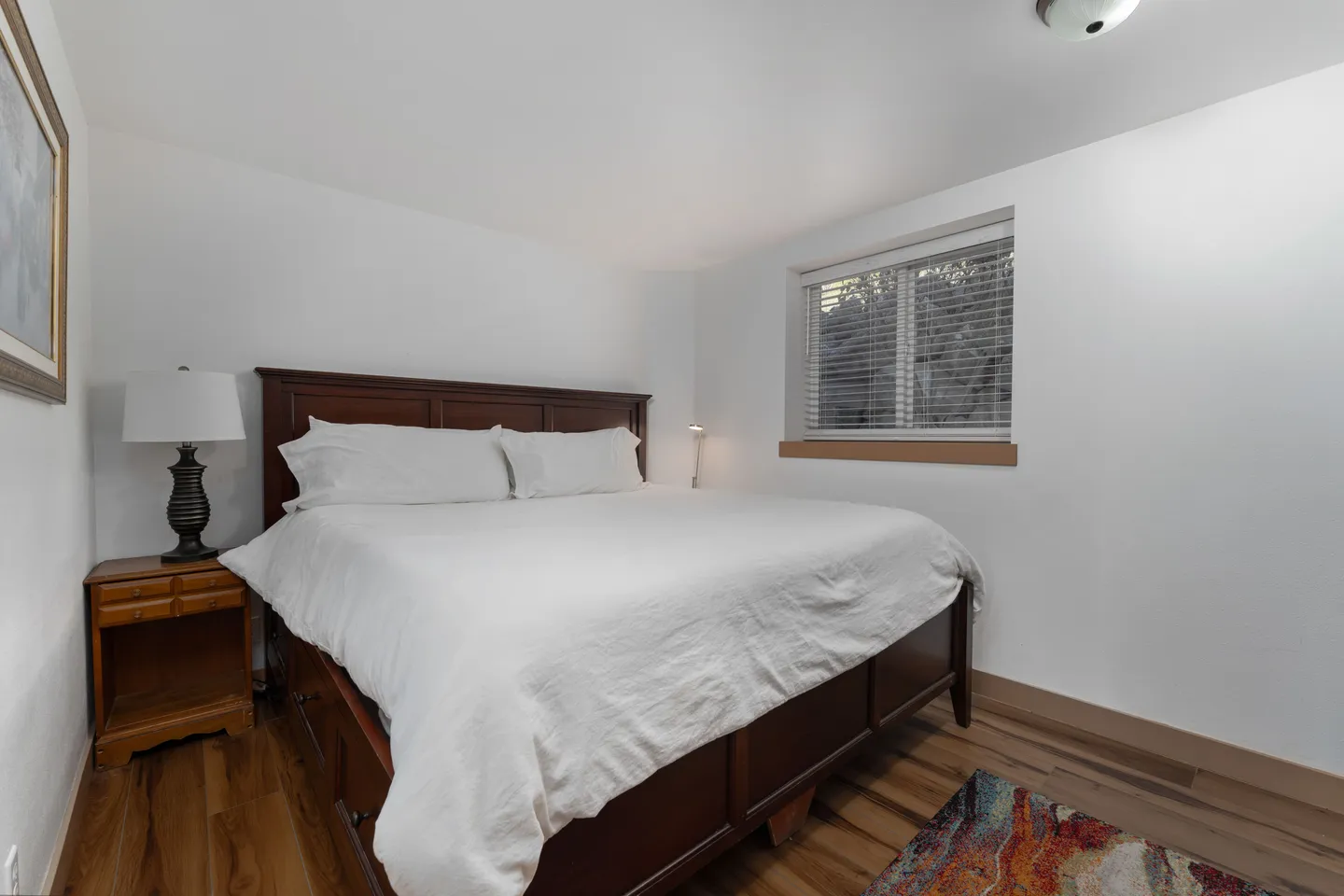 Bedroom featuring a dark wood bed with white bedding, a wooden nightstand with a lamp, and a window with closed blinds.