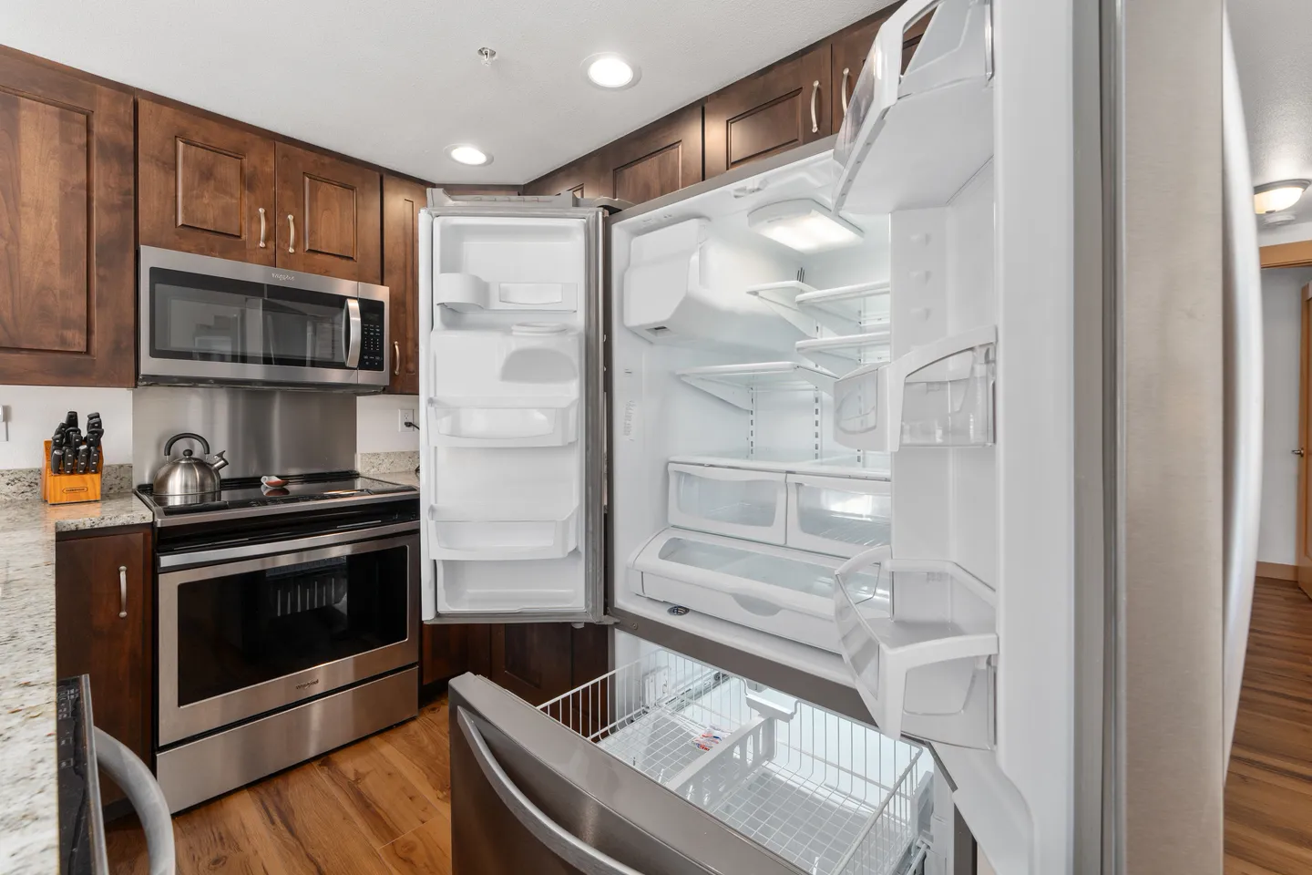 Open stainless steel refrigerator showing its clean interior compartments next to dark wood cabinets, a microwave, and a stove.