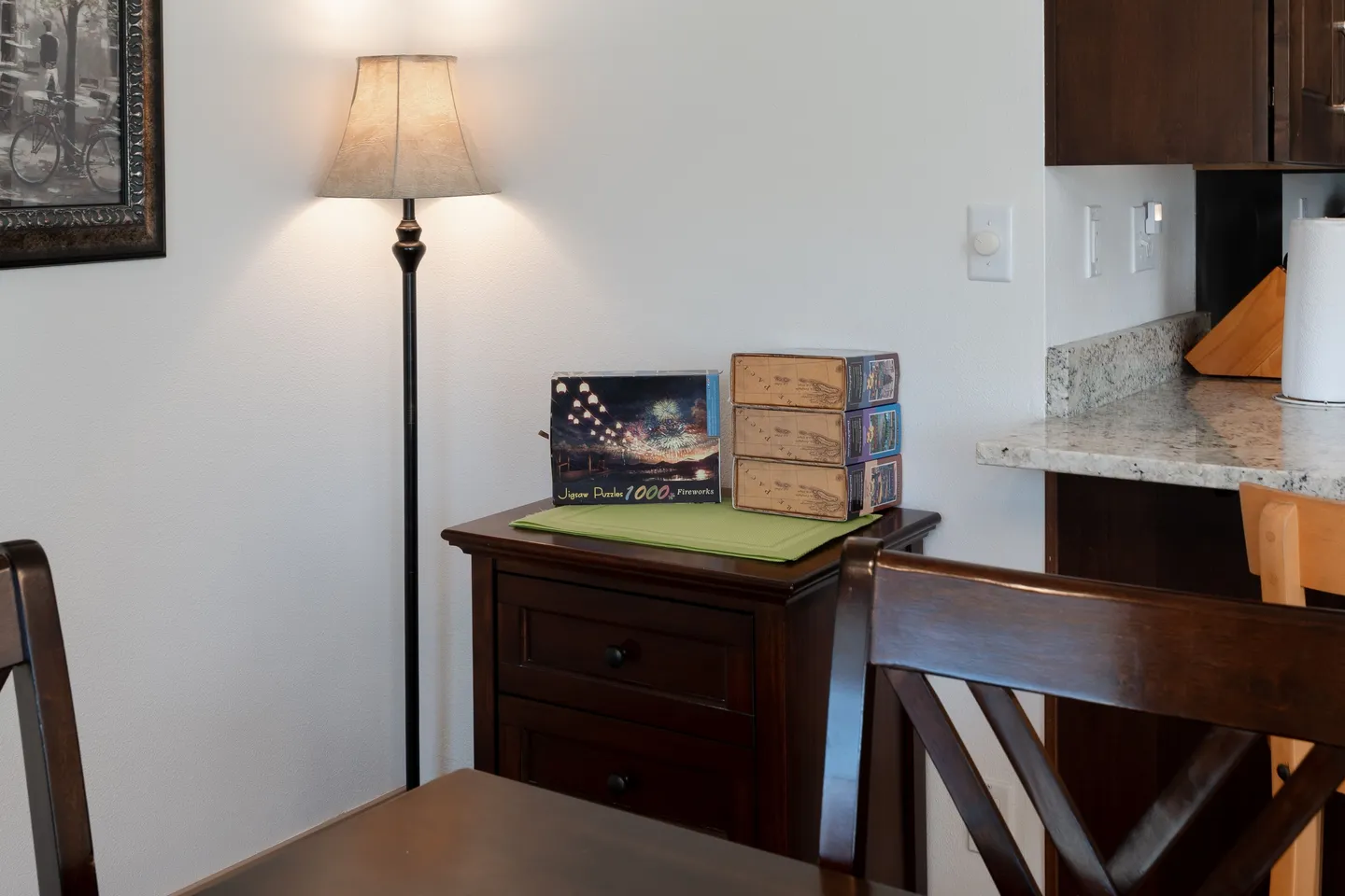 A corner of a room featuring a wooden nightstand with board games and a lit floor lamp next to a kitchen counter.