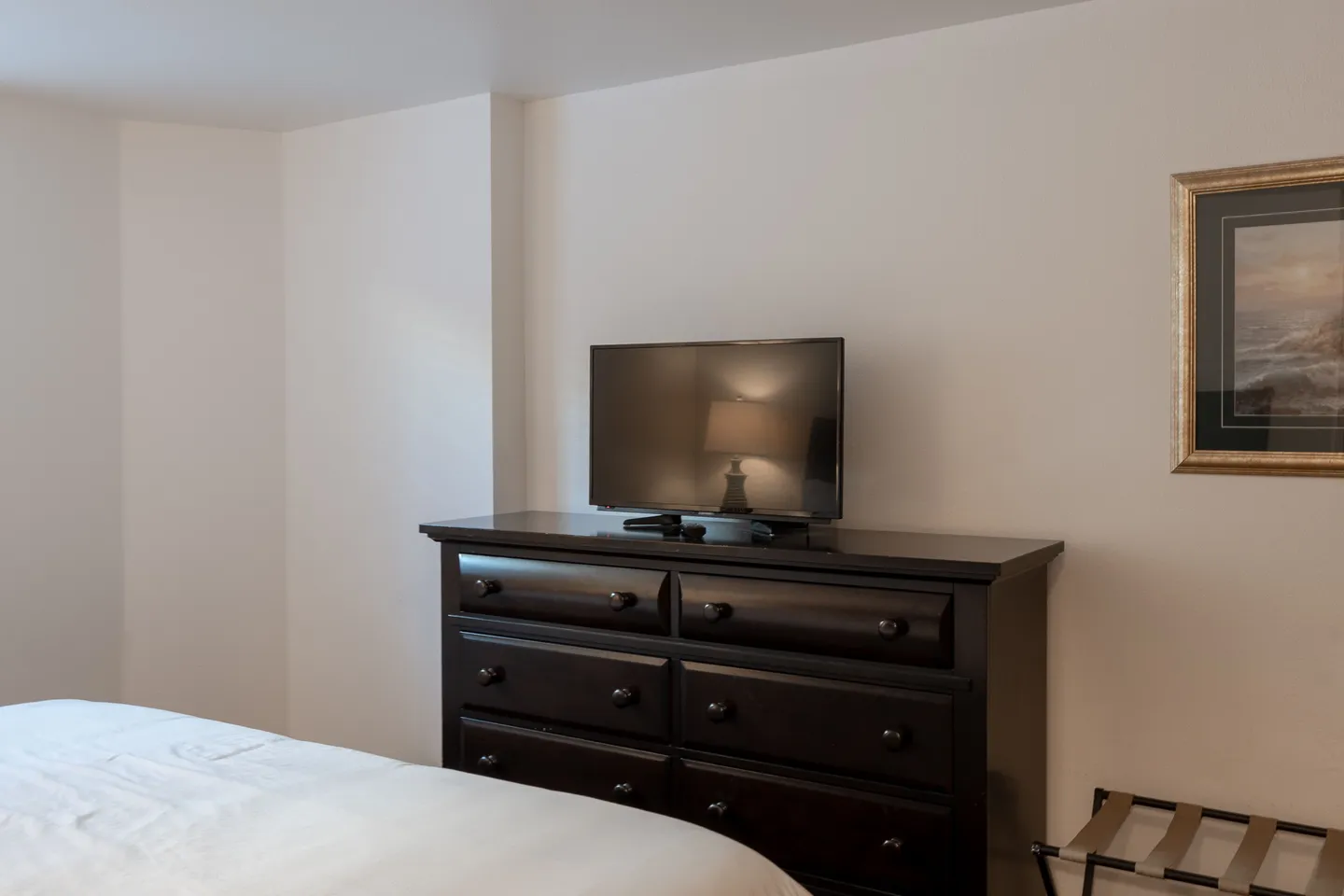 Bedroom featuring a dark wood dresser with a flatscreen TV on top beside a bed and a folding luggage rack.