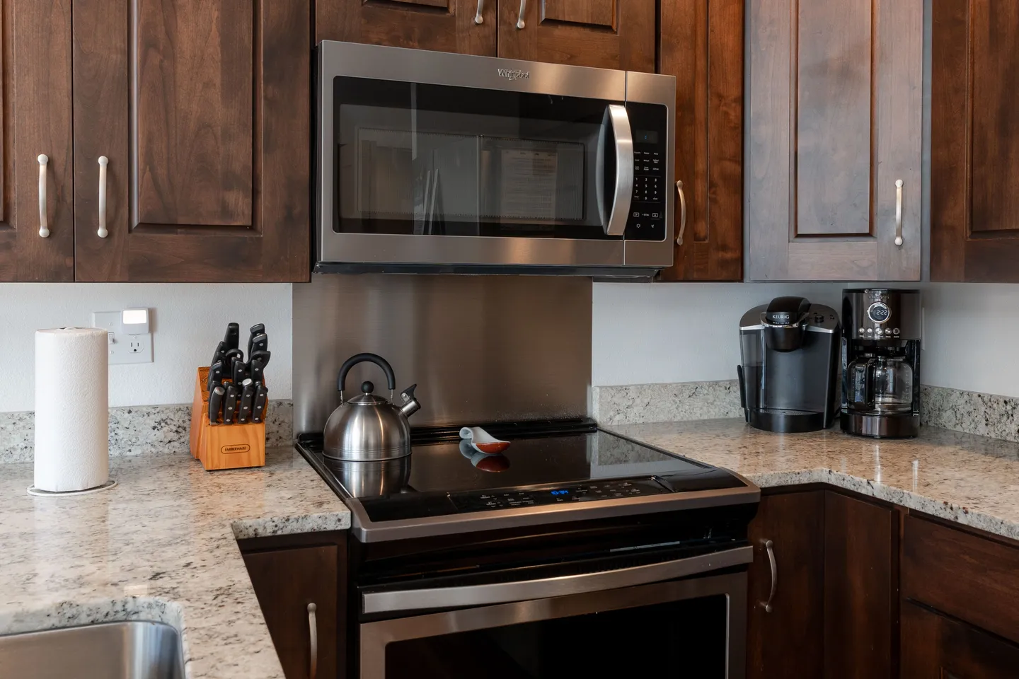 Kitchen counter showing a stainless steel stovetop, overhead microwave, dark wood cabinets, and small appliances.