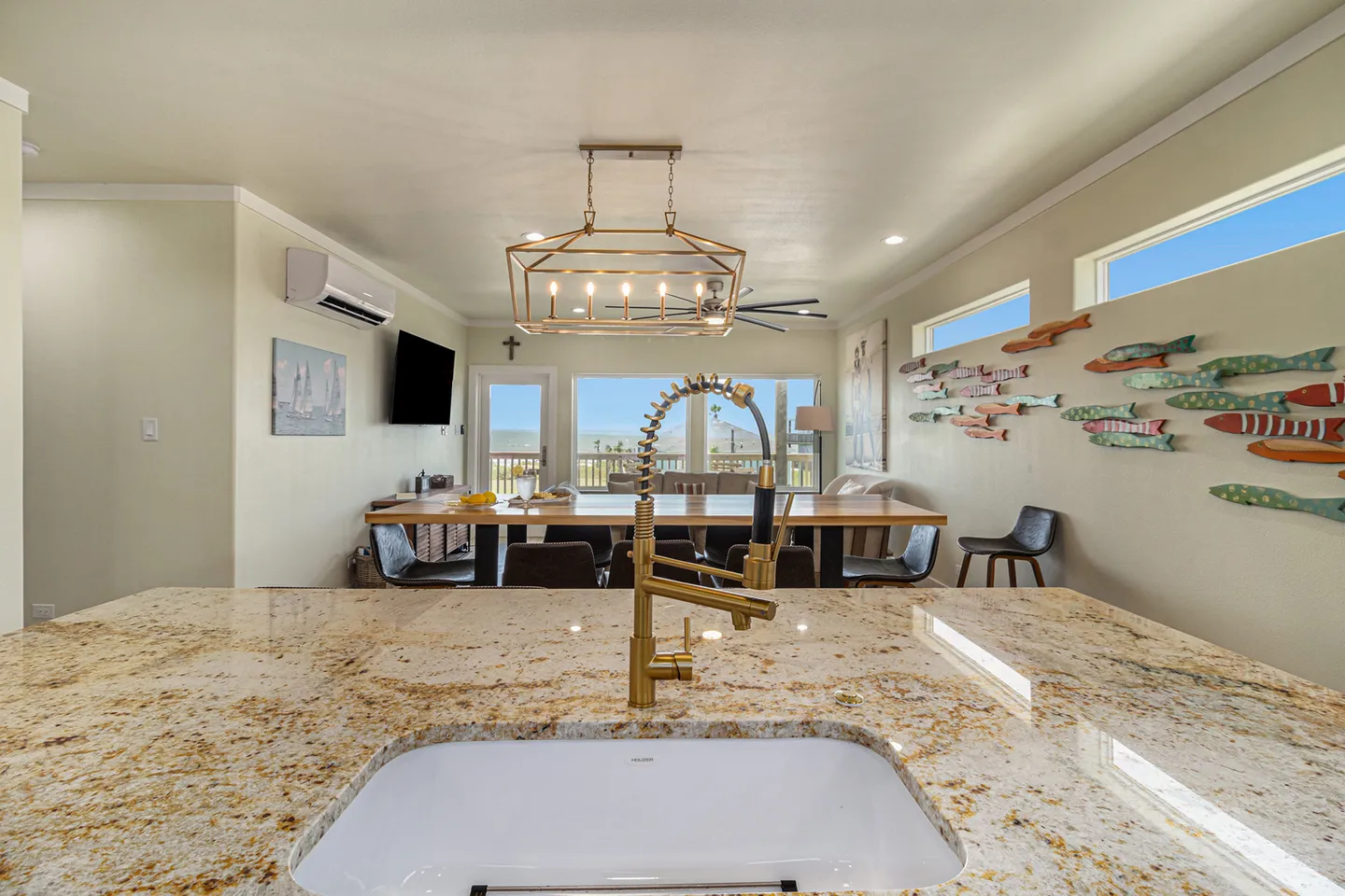 View from an undermount kitchen sink and countertop overlooking an open dining area and windows with an outdoor view.