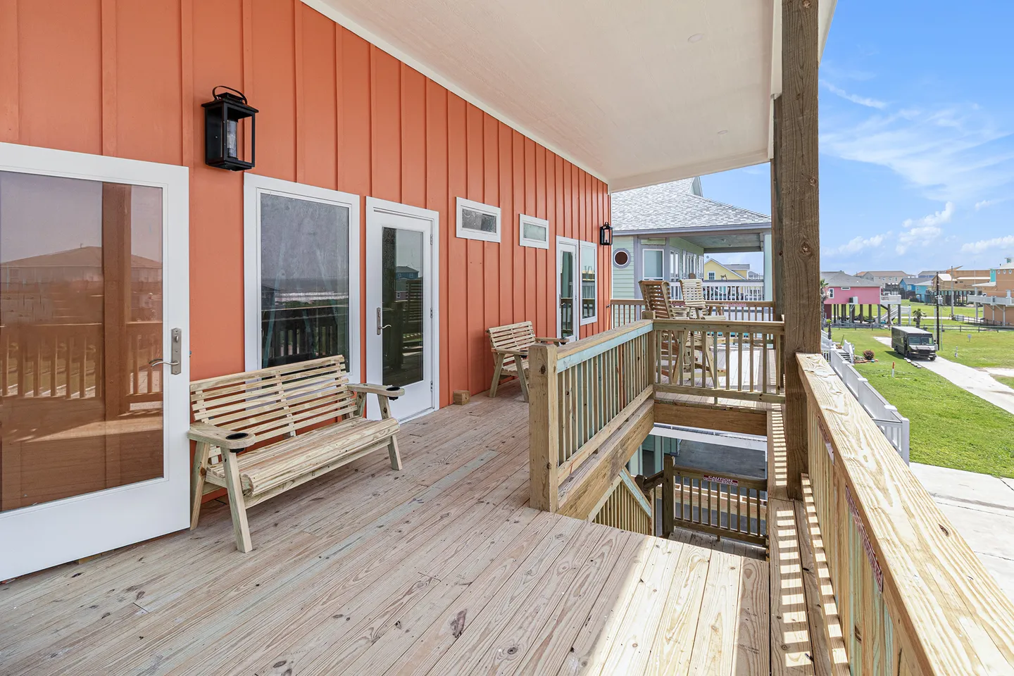 A covered wooden porch along an exterior coral-orange wall with wooden benches, railings, and outdoor stairs leading down.