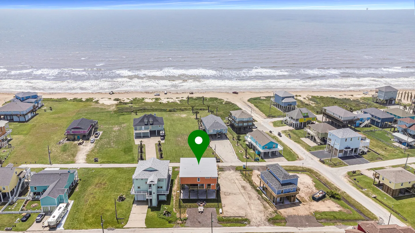 Aerial view of a coastal neighborhood with stilt houses near the sandy beach and ocean, featuring a green location pin over one home.