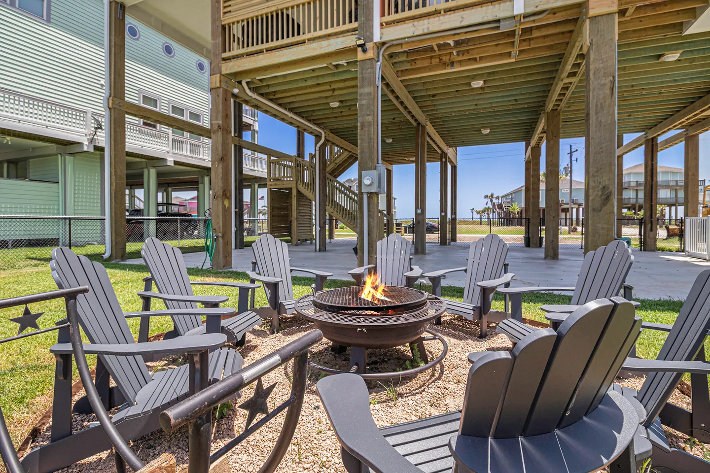 Outdoor fire pit with flames burning, encircled by gray Adirondack chairs in a yard beneath an elevated beach house.