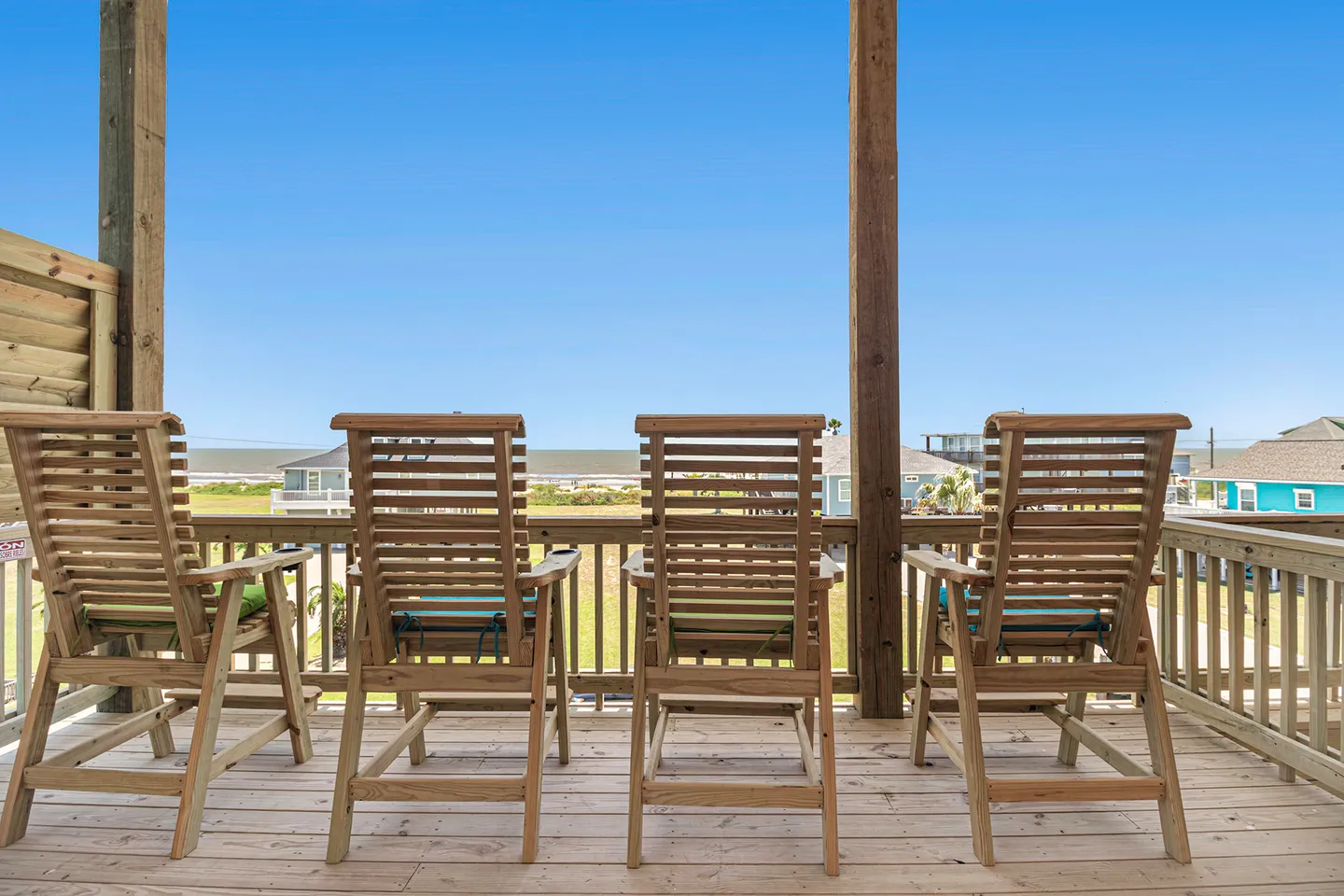 A row of four wooden lounge chairs on a covered wooden deck facing the ocean under a clear blue sky.