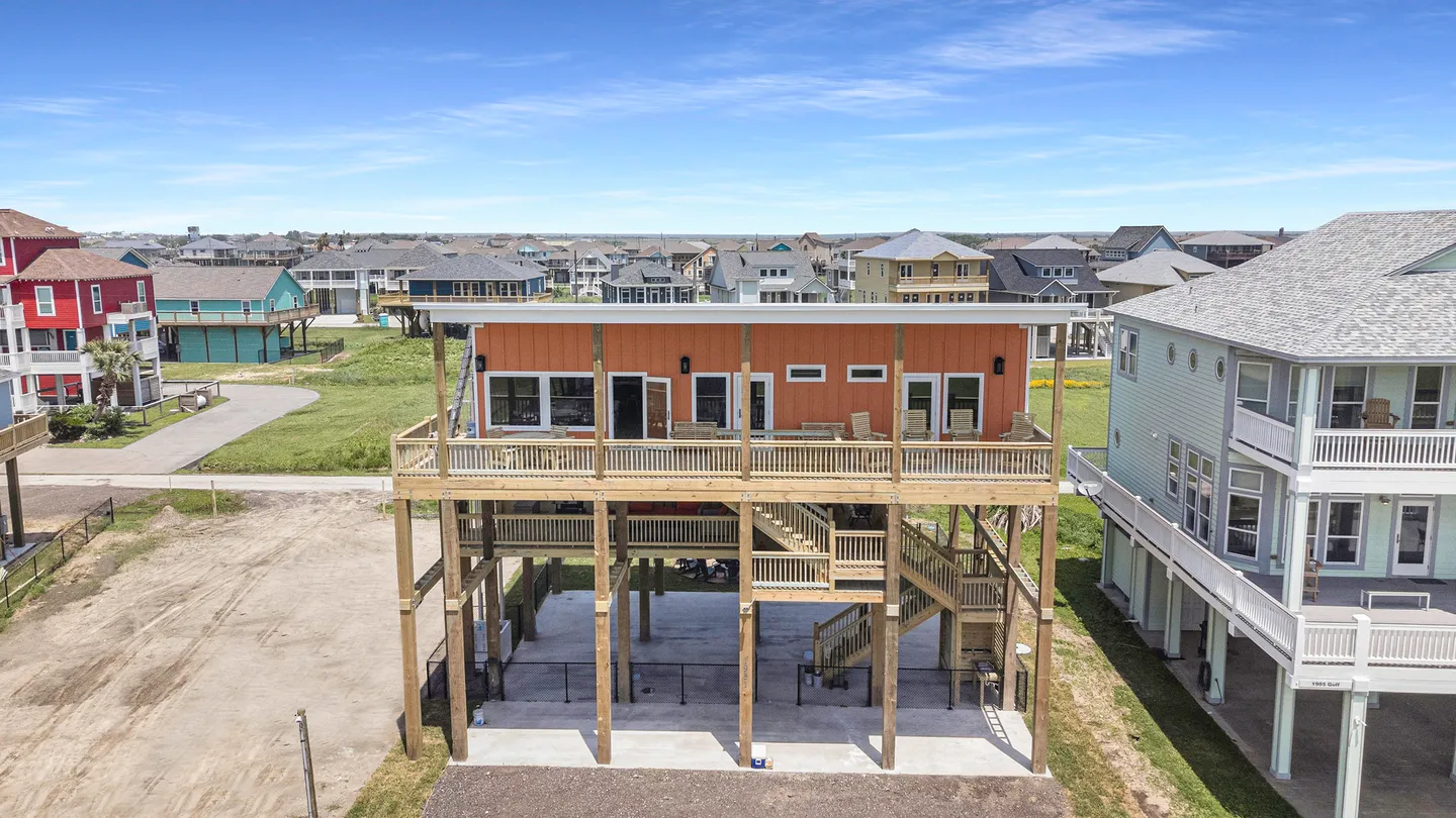 Elevated stilt house with orange siding, a wide wooden deck, outdoor stairs, and a neighborhood view under a blue sky.