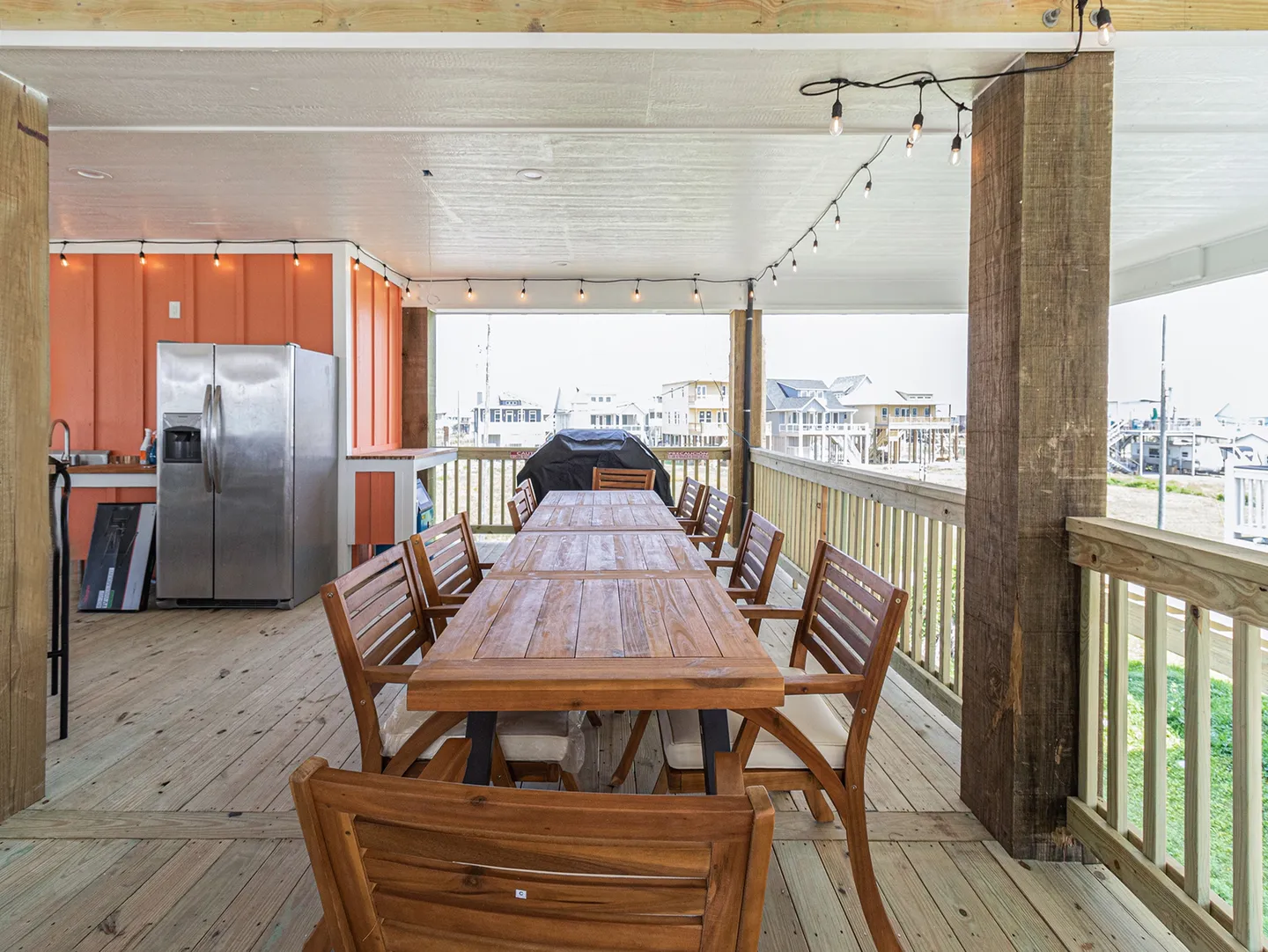 Covered outdoor deck with a long wooden dining table, chairs, a refrigerator, and a barbecue grill overlooking surrounding houses.