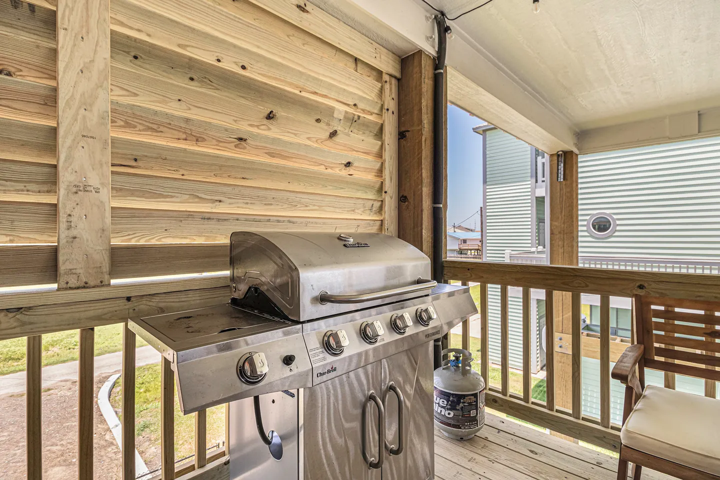 A stainless steel gas grill on a covered wooden deck with a wooden chair next to the railing.