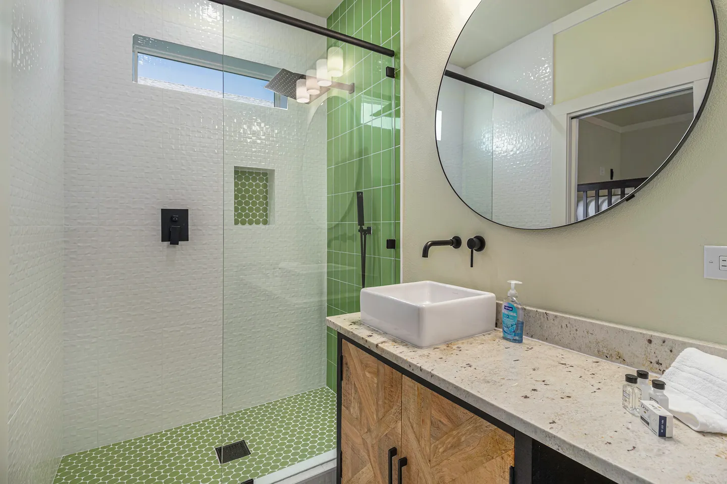 Bathroom with a glass walk-in shower featuring green tile, a vessel sink on a speckled countertop, and a round wall mirror.