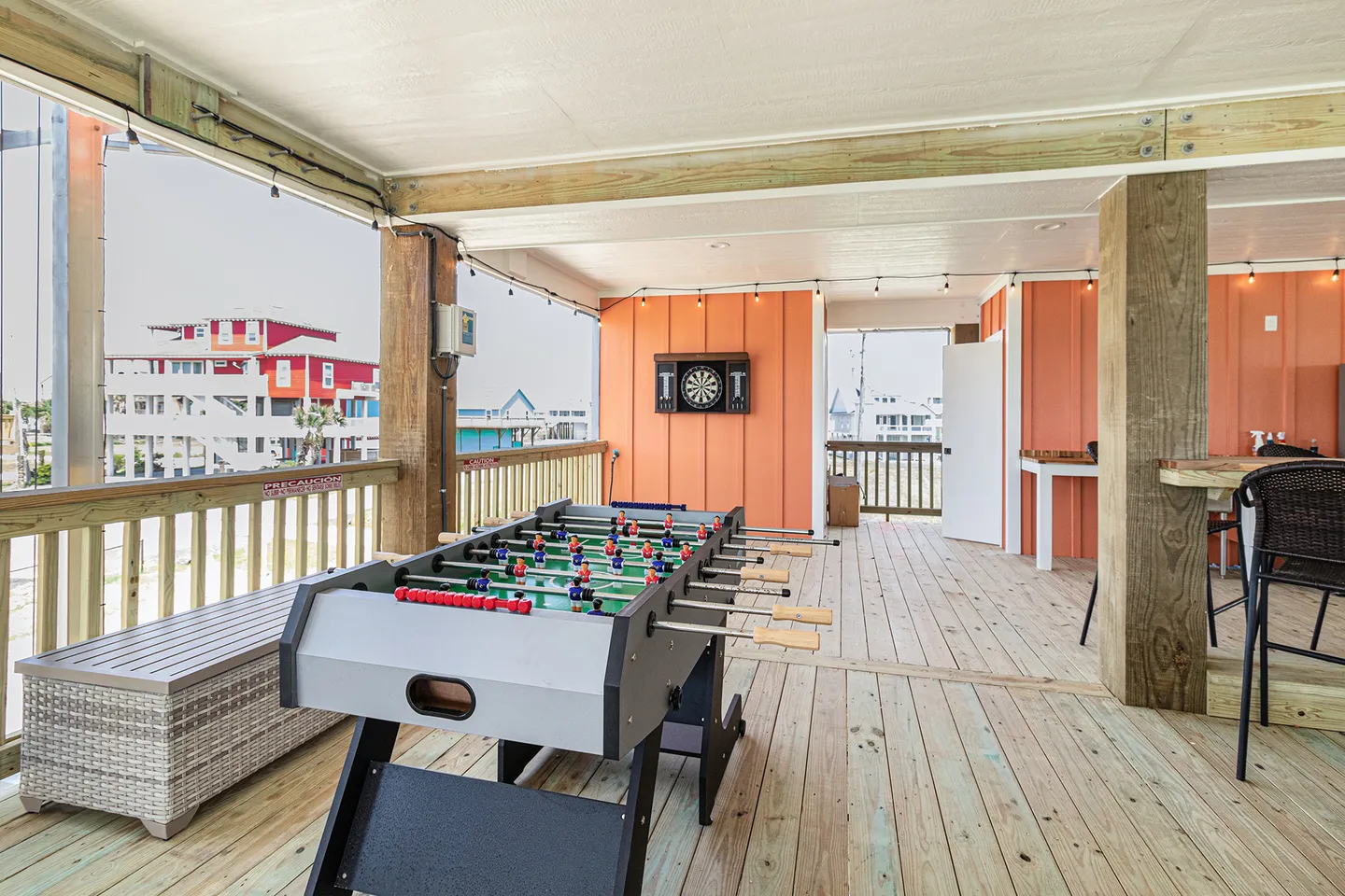 A covered outdoor deck featuring a foosball table, a dartboard mounted on an orange wall, and outdoor bar seating.