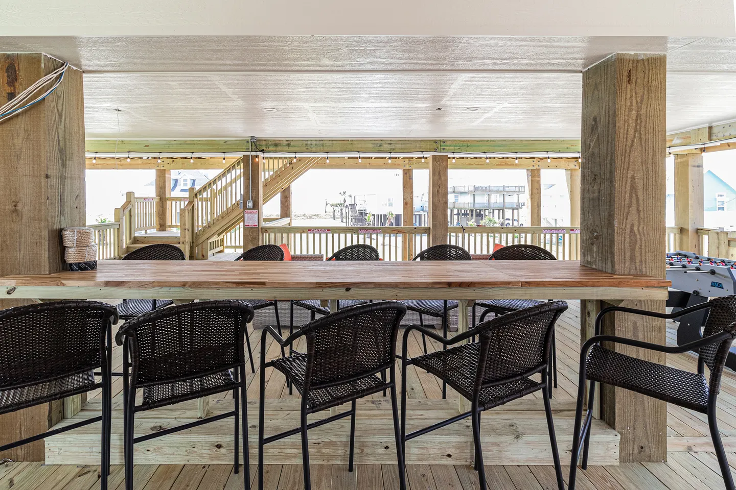Covered outdoor patio under a stilt house with a built-in wooden bar counter, wicker armchairs, and a foosball table.