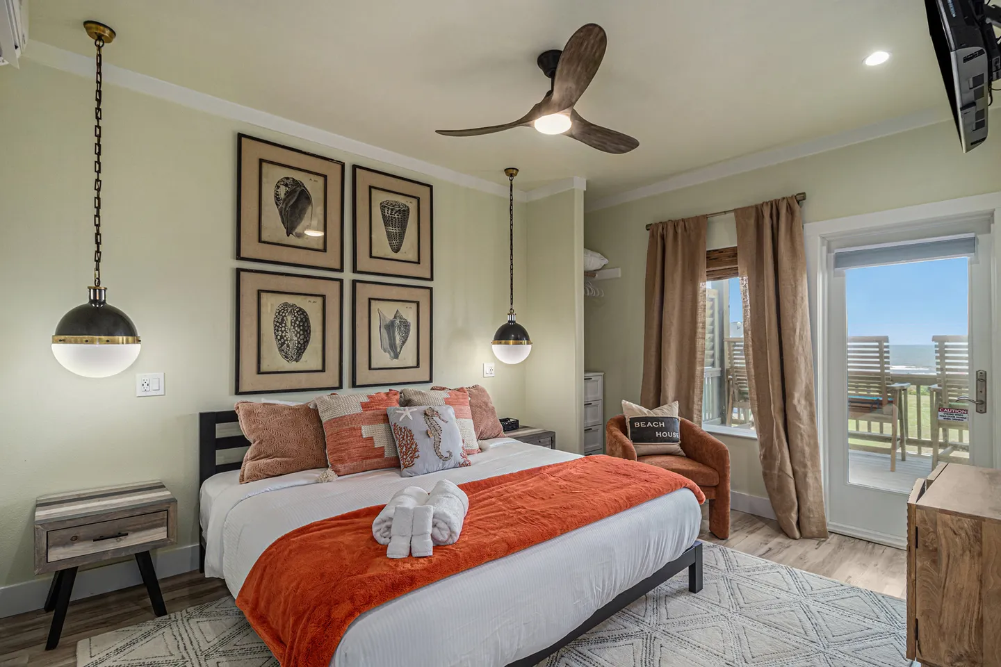 Bedroom with a large bed dressed in white and orange linens, framed shell art on the wall, pendant lights, and a door opening to an outdoor patio with ocean views.