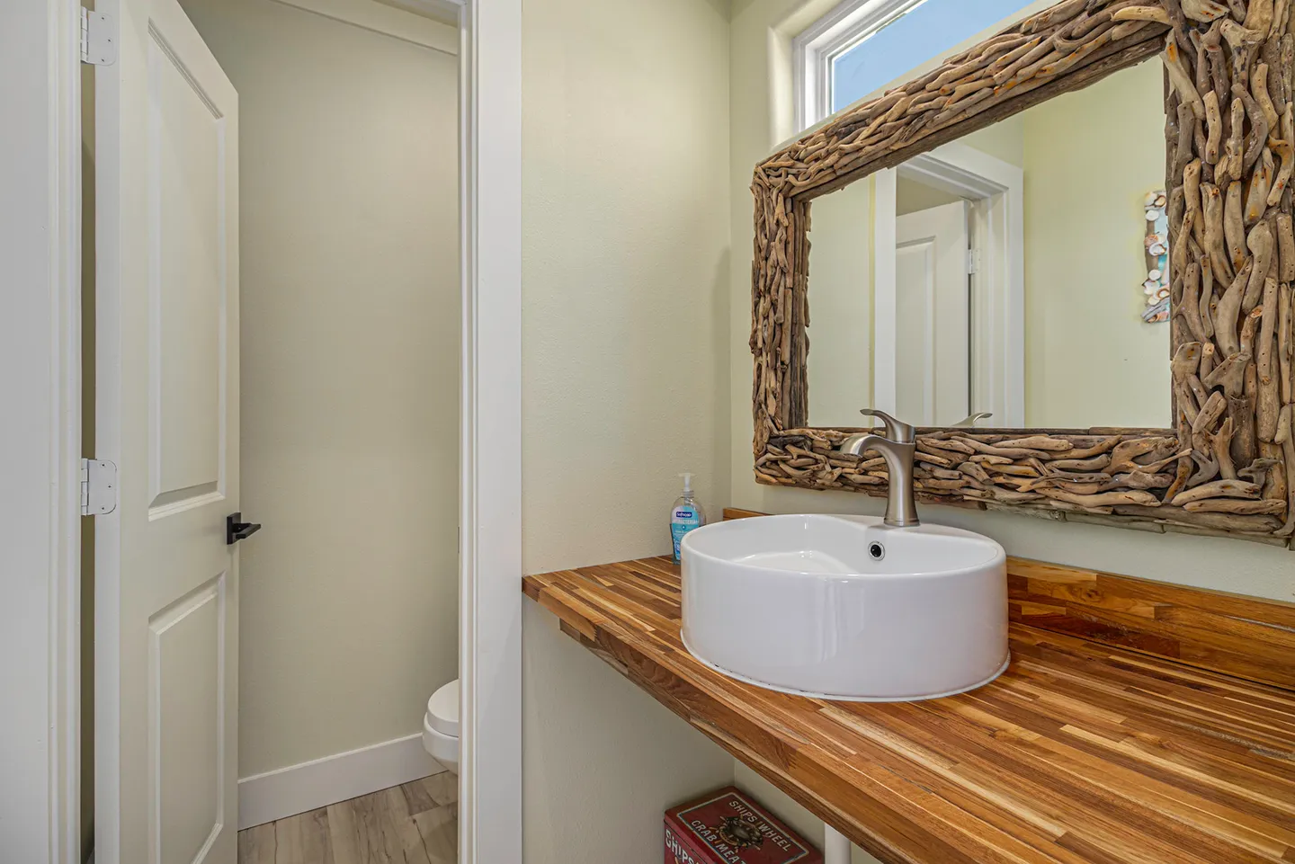 Powder room vanity featuring a vessel sink on a butcher-block counter beneath a driftwood-framed mirror, next to an open doorway to the toilet.
