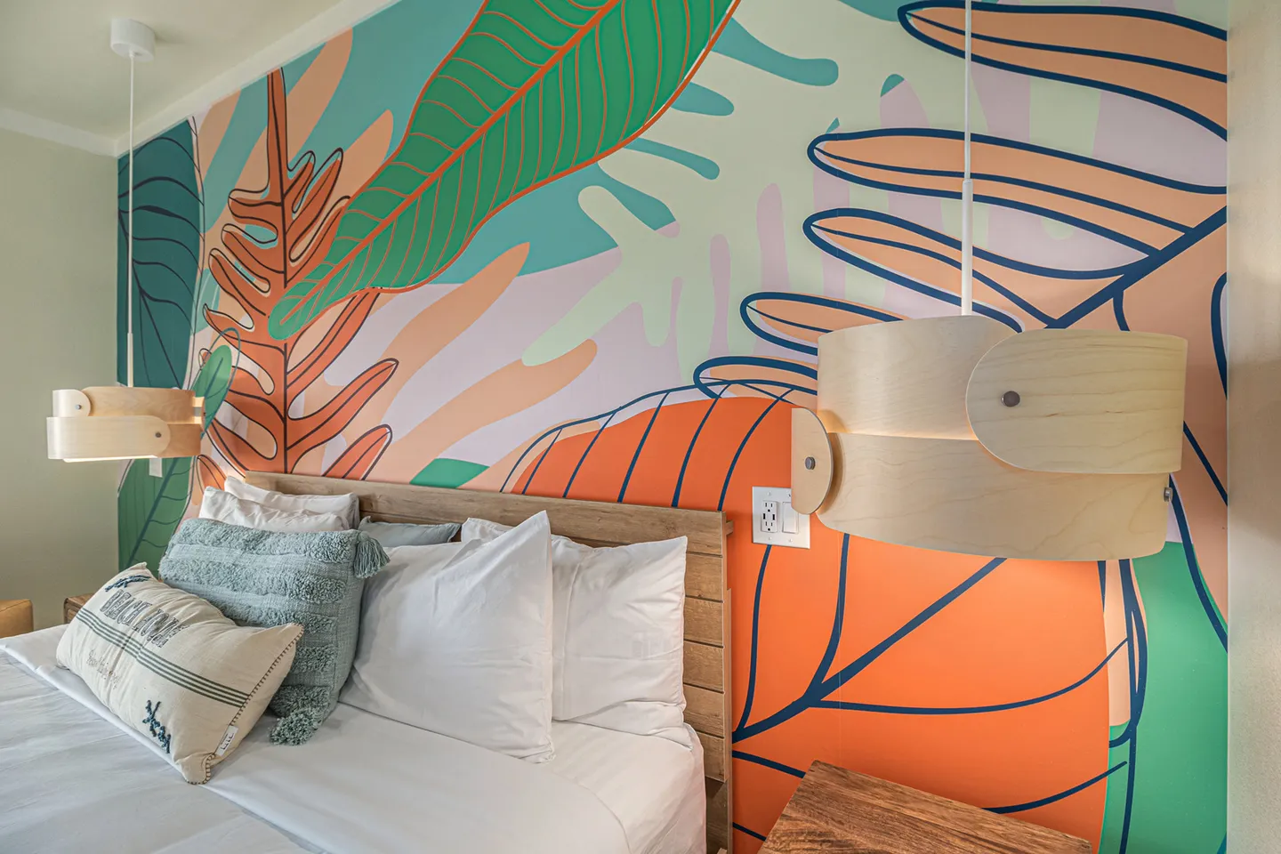Bedroom featuring a colorful tropical leaf wall mural behind a wooden headboard, made bed, and hanging wood pendant lights.