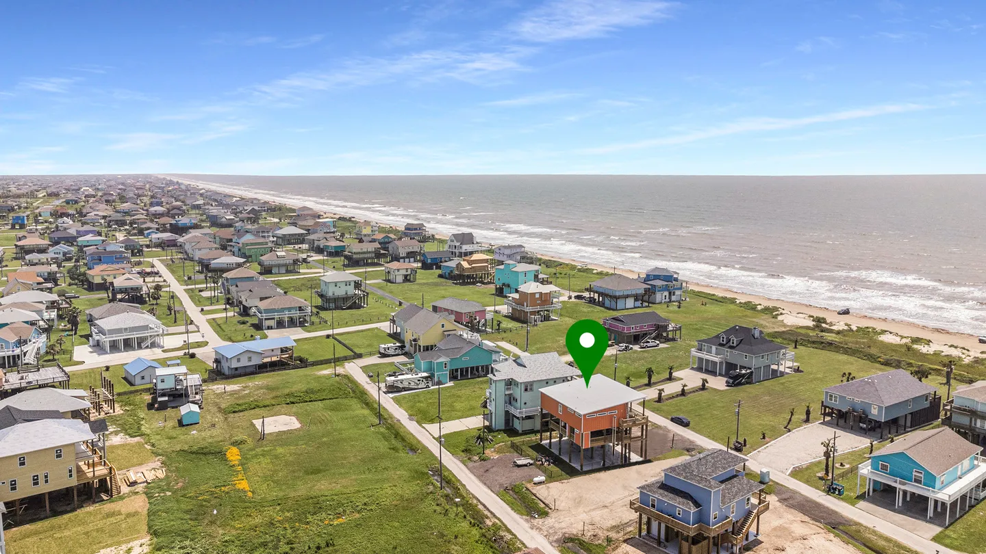 Aerial view of a coastal neighborhood with stilt homes near the beach, highlighting an orange and white elevated house with a green map pin.
